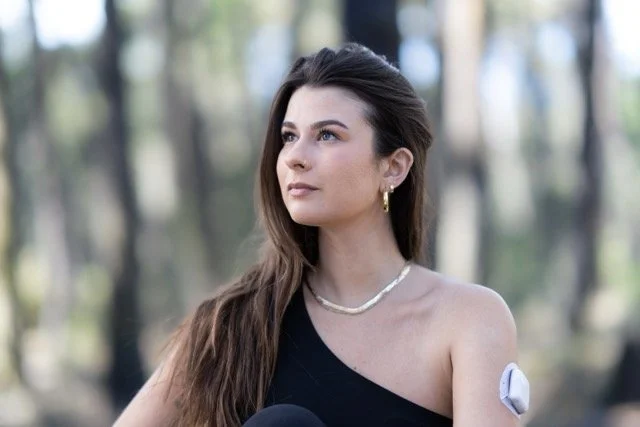 A young woman with long brown hair, wearing a black off-shoulder top, gold earrings, and a pearl necklace, standing outdoors in a forested area.