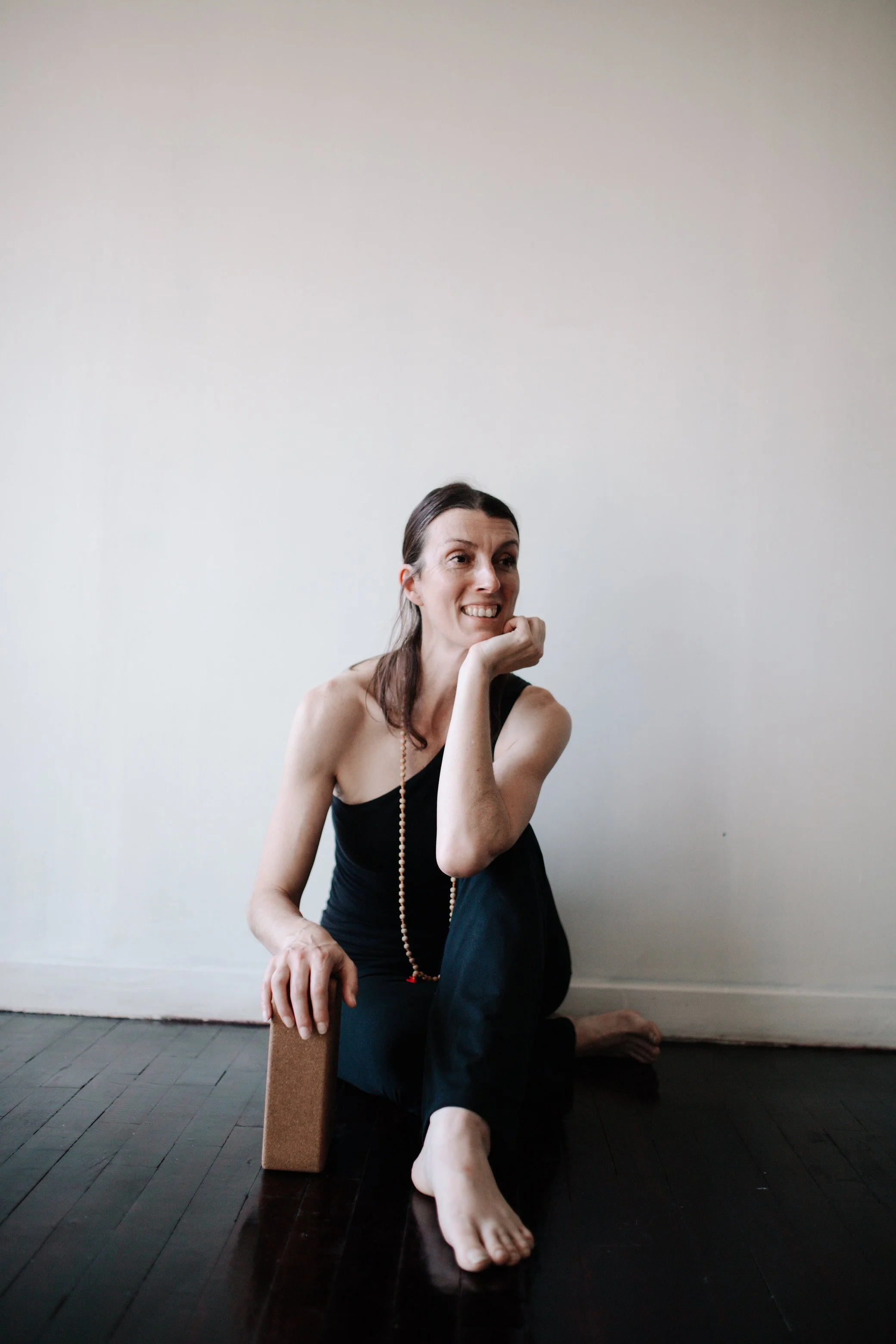 A woman with long brown hair and light skin sitting on a dark wooden floor, resting her left arm on a cork yoga block, and smiling while looking to her right against a plain white wall. She wears a black tank top, black pants, and a beaded necklace.