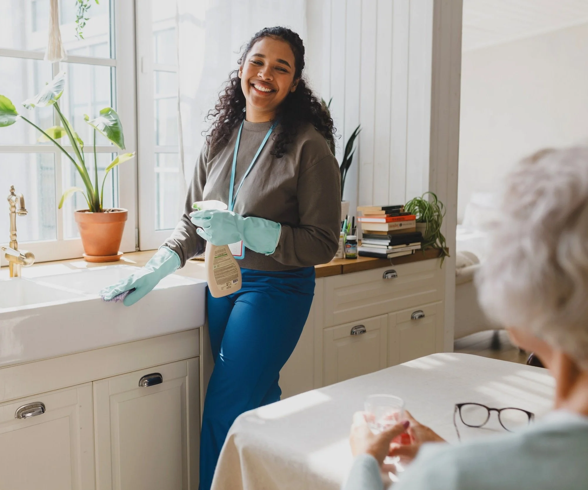 Young carer smiling while cleaning a kitchen counter with gloves and spray bottle, supporting an elderly woman at home, representing domestic assistance and personalised home care services.