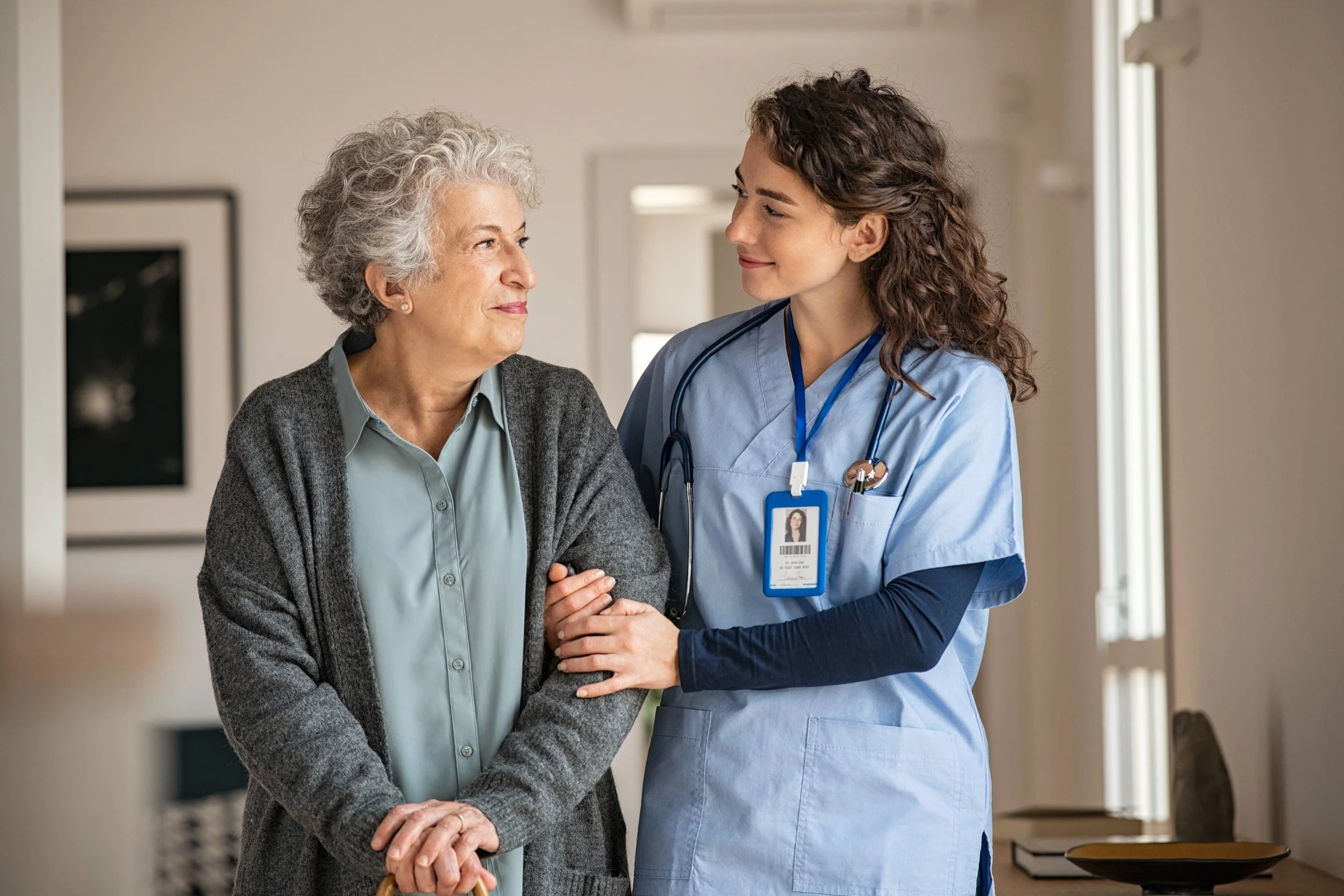 Healthcare professional in blue scrubs with stethoscope supporting an elderly woman with silver hair, symbolising compassionate in-home nursing, aged care and health monitoring.