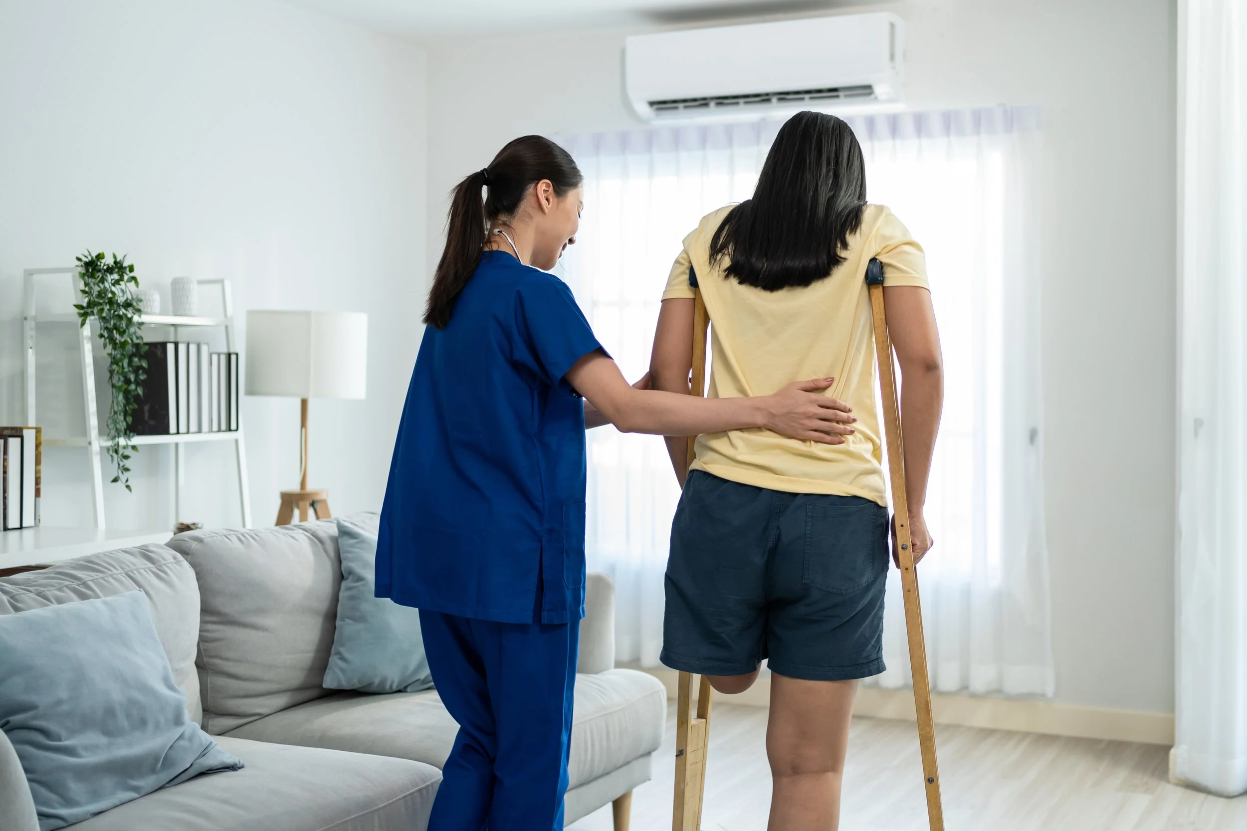 Healthcare worker supporting a woman with a leg injury as she walks with crutches in a bright home environment, representing personalised rehabilitation, in-home nursing and mobility support.