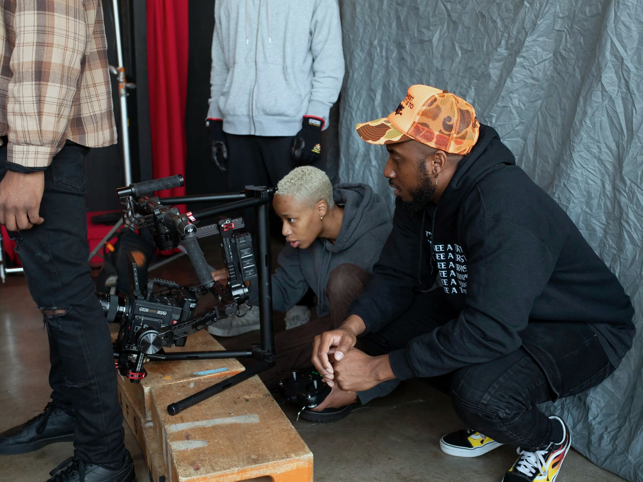 Elton Holt and Ariel K Harris  working on setting up a camera rig for filming. One is crouching and adjusting the equipment, the other is sitting and observing the camera, with a third person standing nearby in the background.
