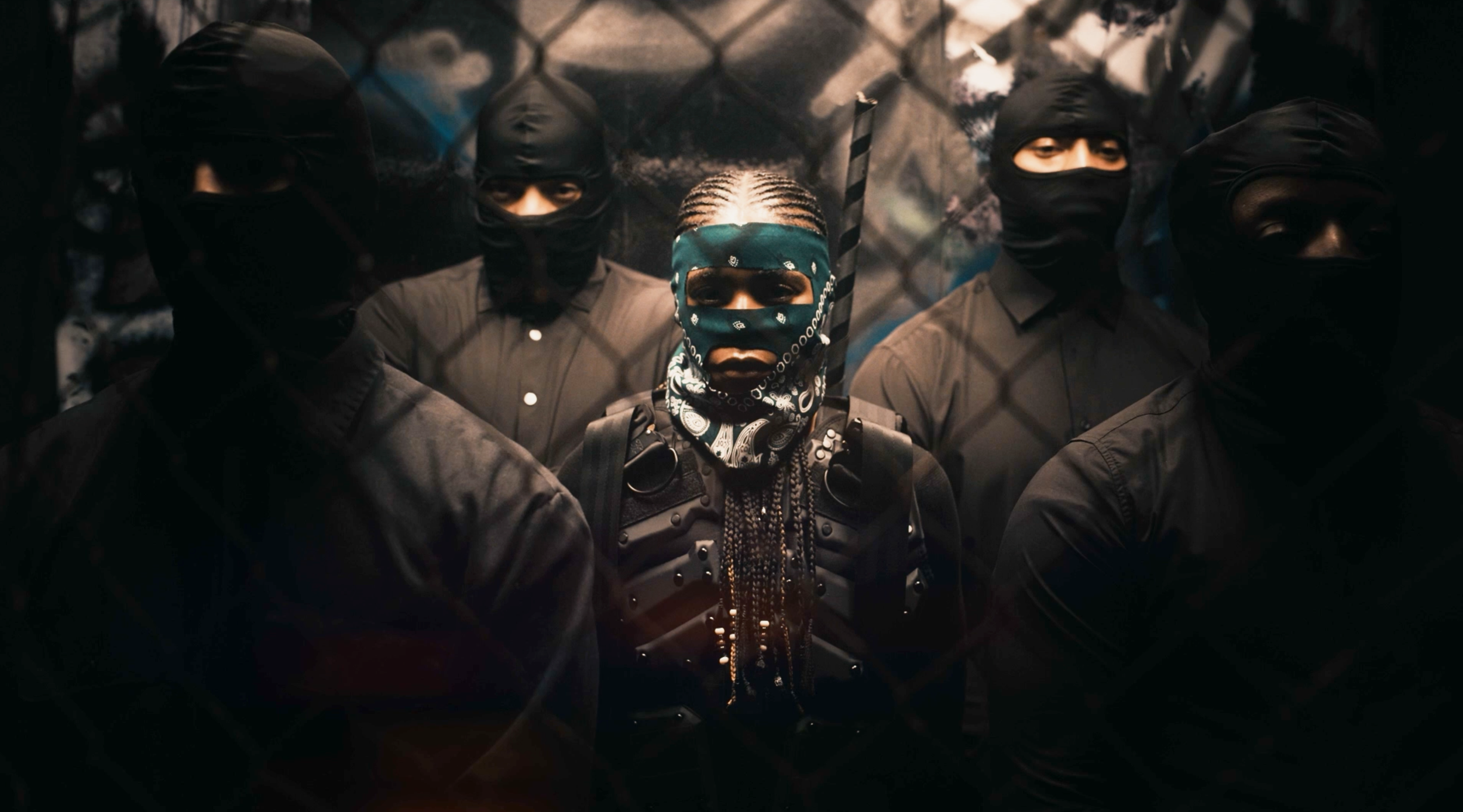A group of five people wearing dark clothing and black masks, standing behind a chain-link fence, with a woman in the center wearing a bandana covering her face and braided hair.