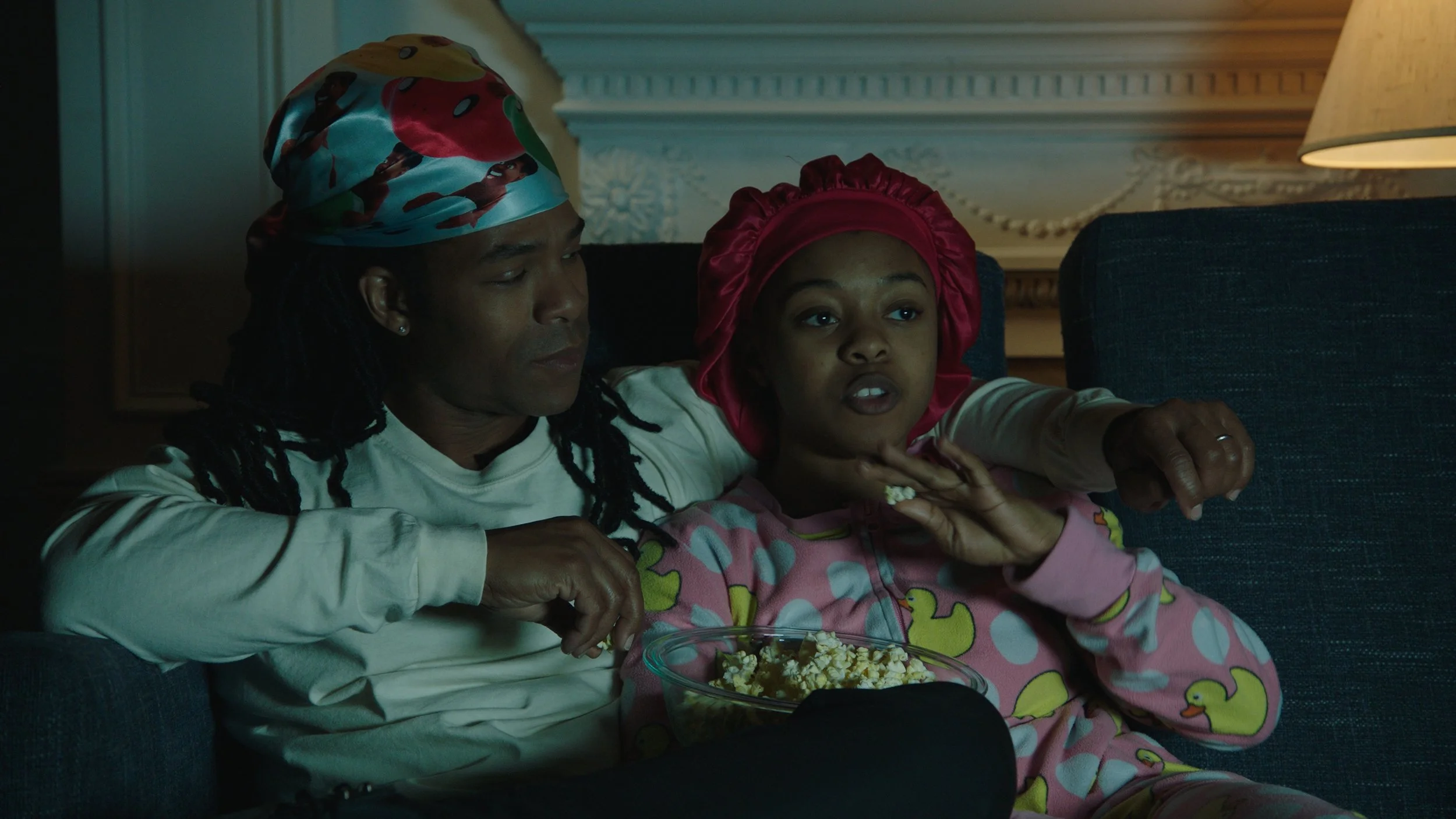 A man and a girl watching TV together on a couch, eating popcorn, with the girl wearing a red bonnet and the man wearing a colorful sleep cap.