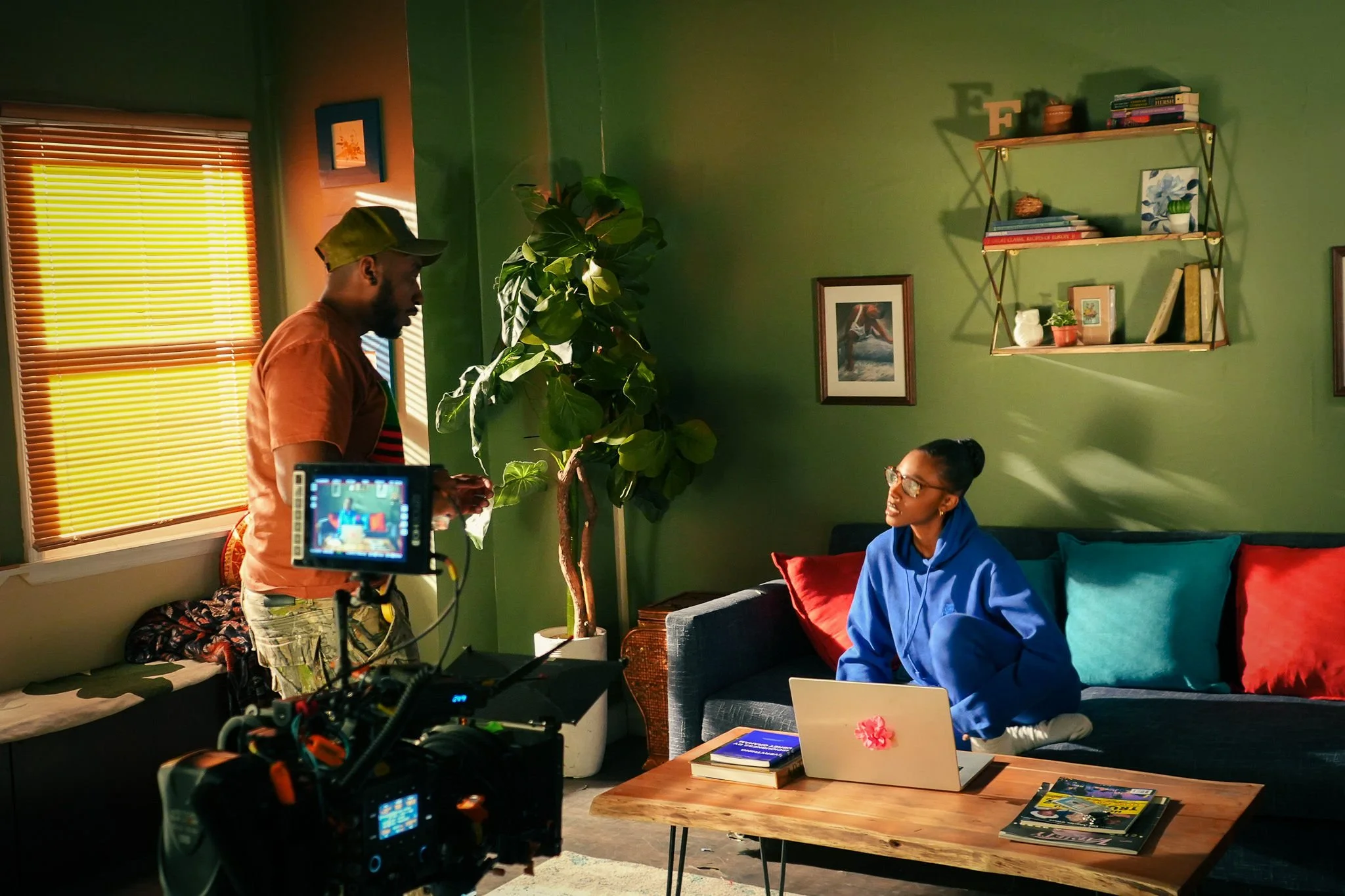 Elton Holt directs Ariana as a camera on a tripod records the scene, and sunlight comes through a window with yellow blinds.