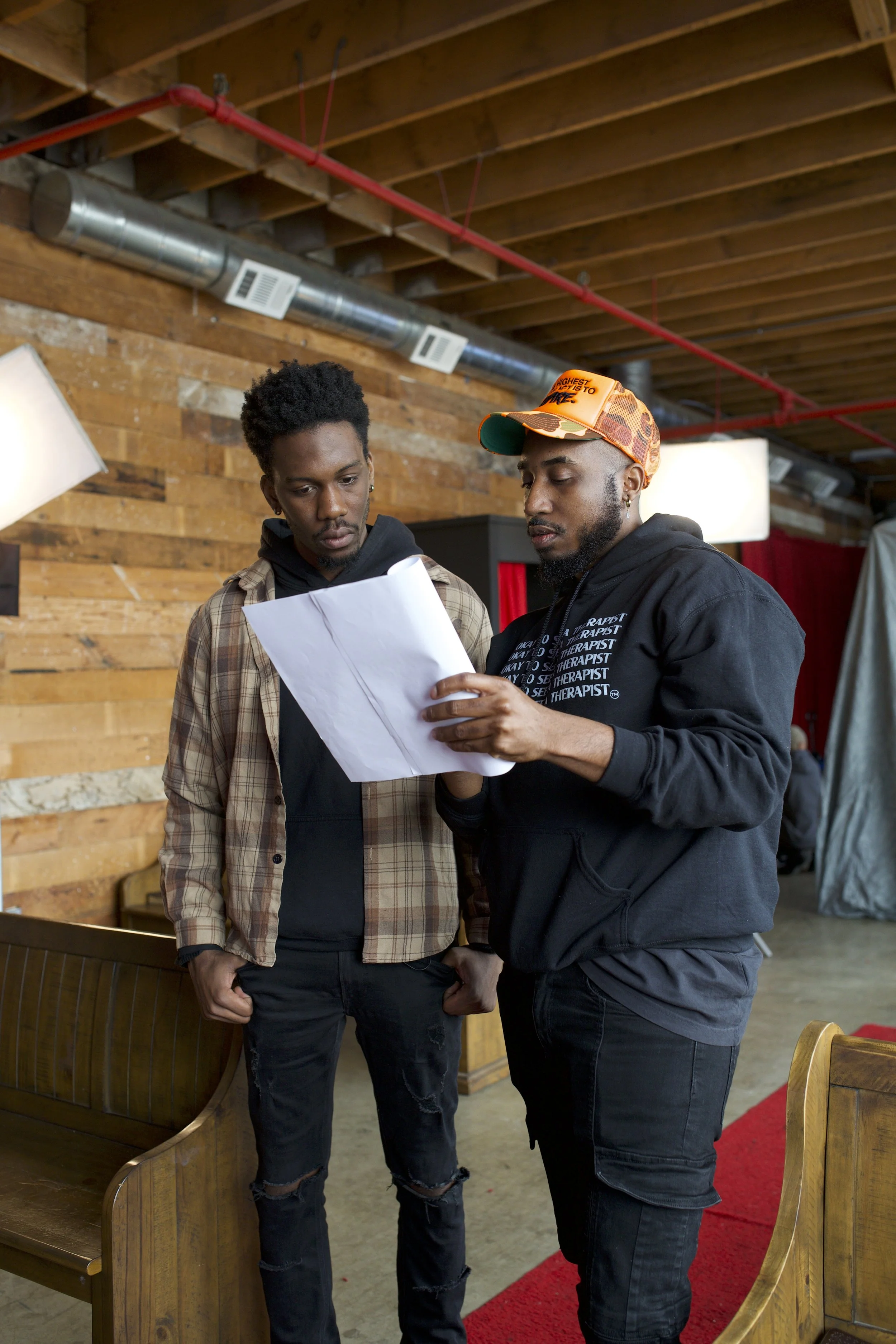 Two men are looking at a piece of paper together indoors, with wood-paneled walls and some lighting fixtures in the background.