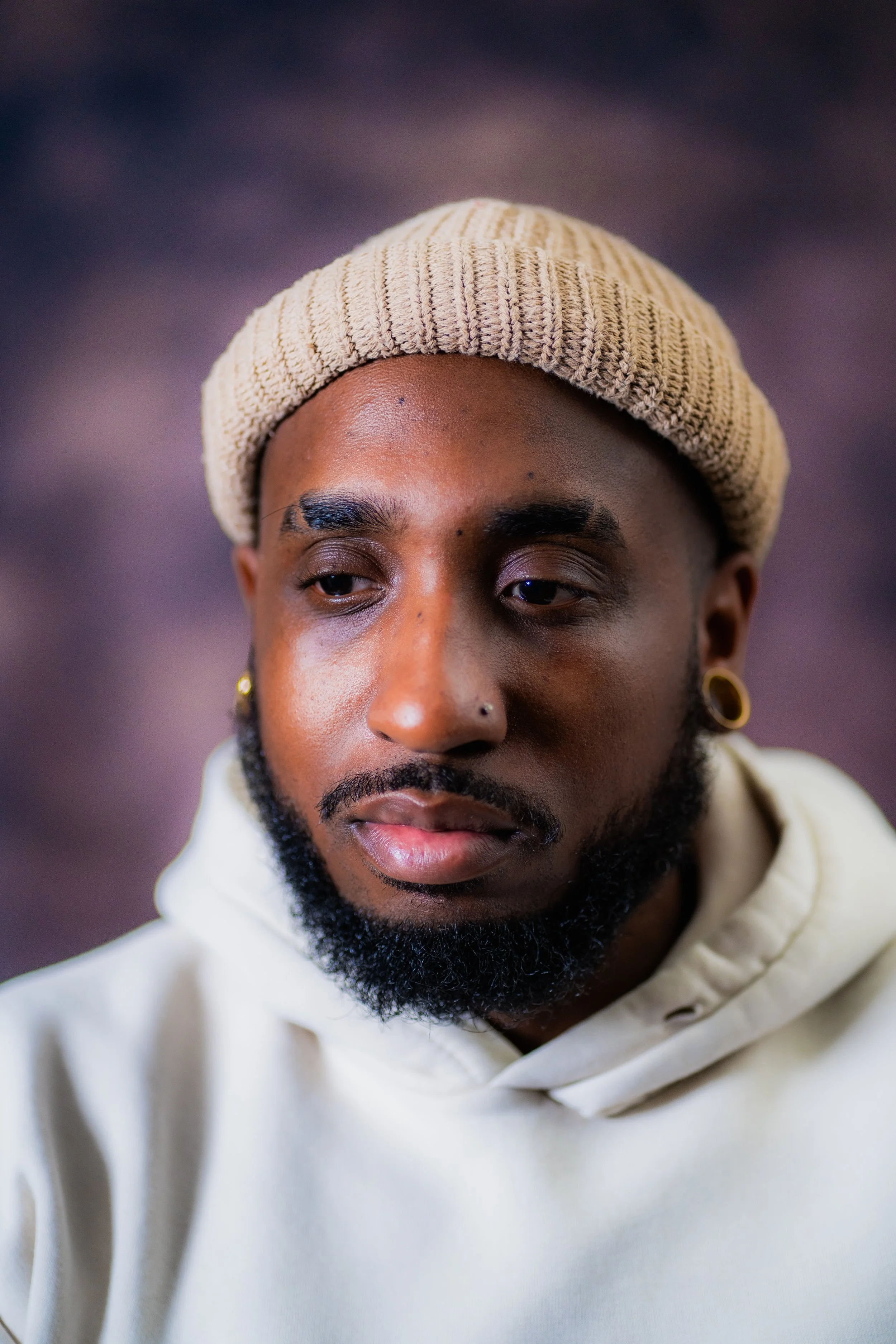 Close-up portrait of a young man with a beard and multiple piercings, wearing a beige knit cap and cream hoodie, with a blurred dark background.