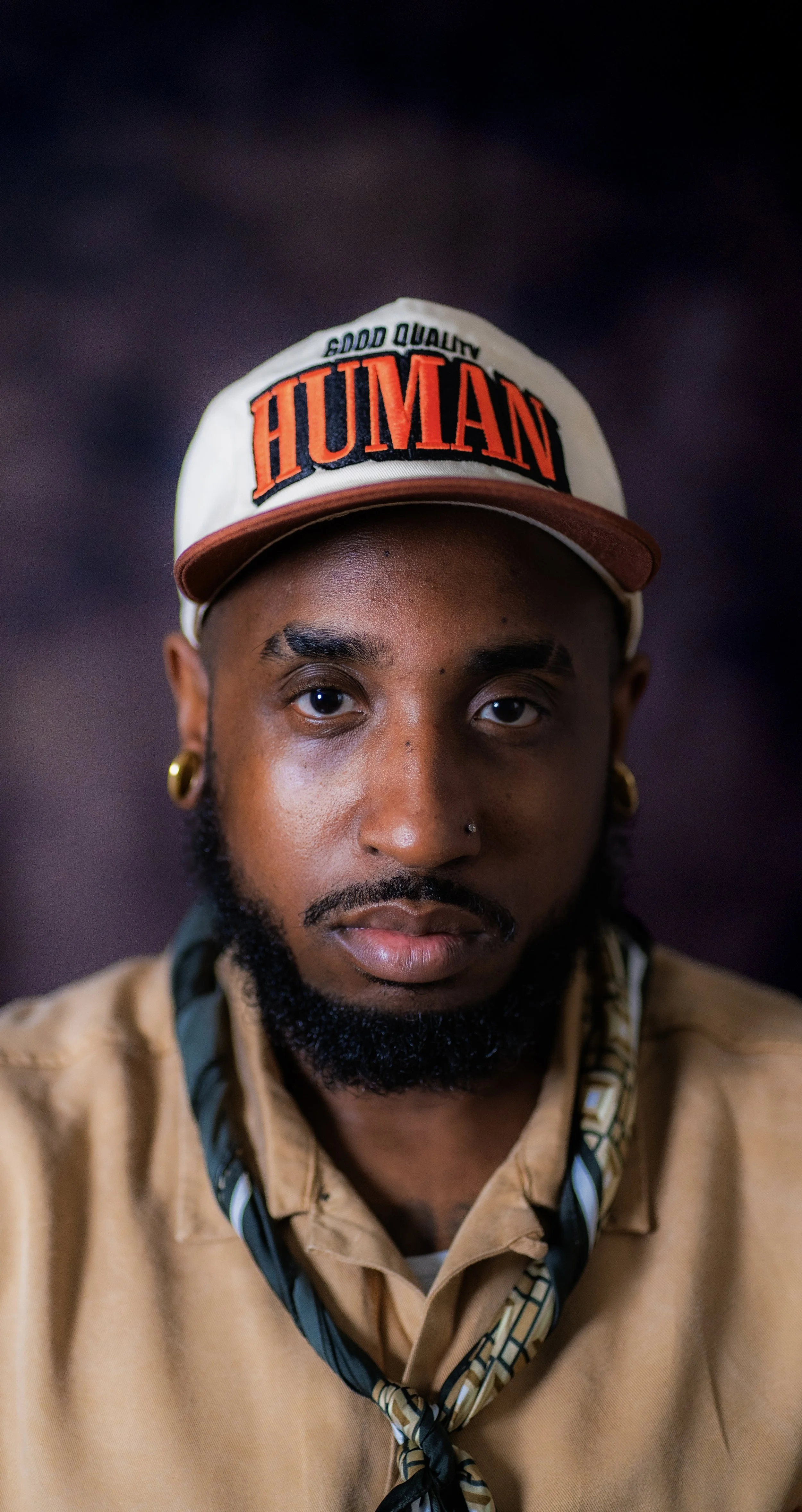 Elton Holt, with a beard, gold hoop earrings, and a nose piercing, wearing a beige shirt, a patterned neck scarf, and a white cap with red and black text that says "HUMAN" and "Good Quality".