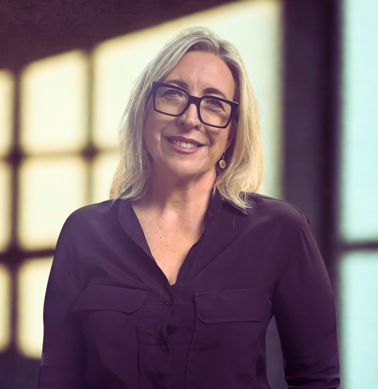 A woman with blonde hair, black glasses, and earrings, smiling in front of a blurred window background.