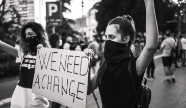 Protesters standing outdoors, one woman holding a sign that reads 'WE NEED A CHANGE,' both women wearing face masks.