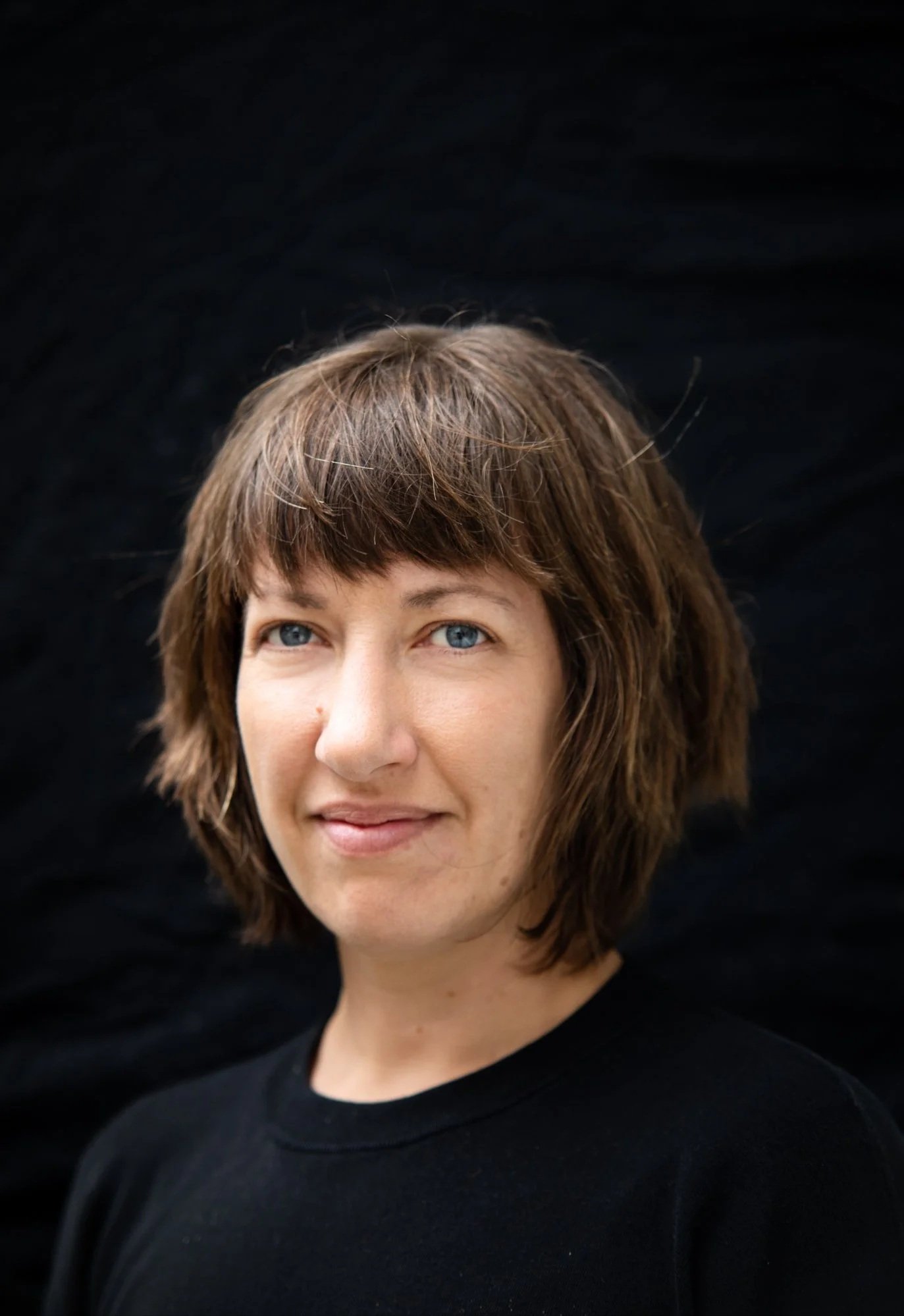A woman with short, brown, slightly wavy hair and blue eyes, wearing a black top, smiles softly against a black background.