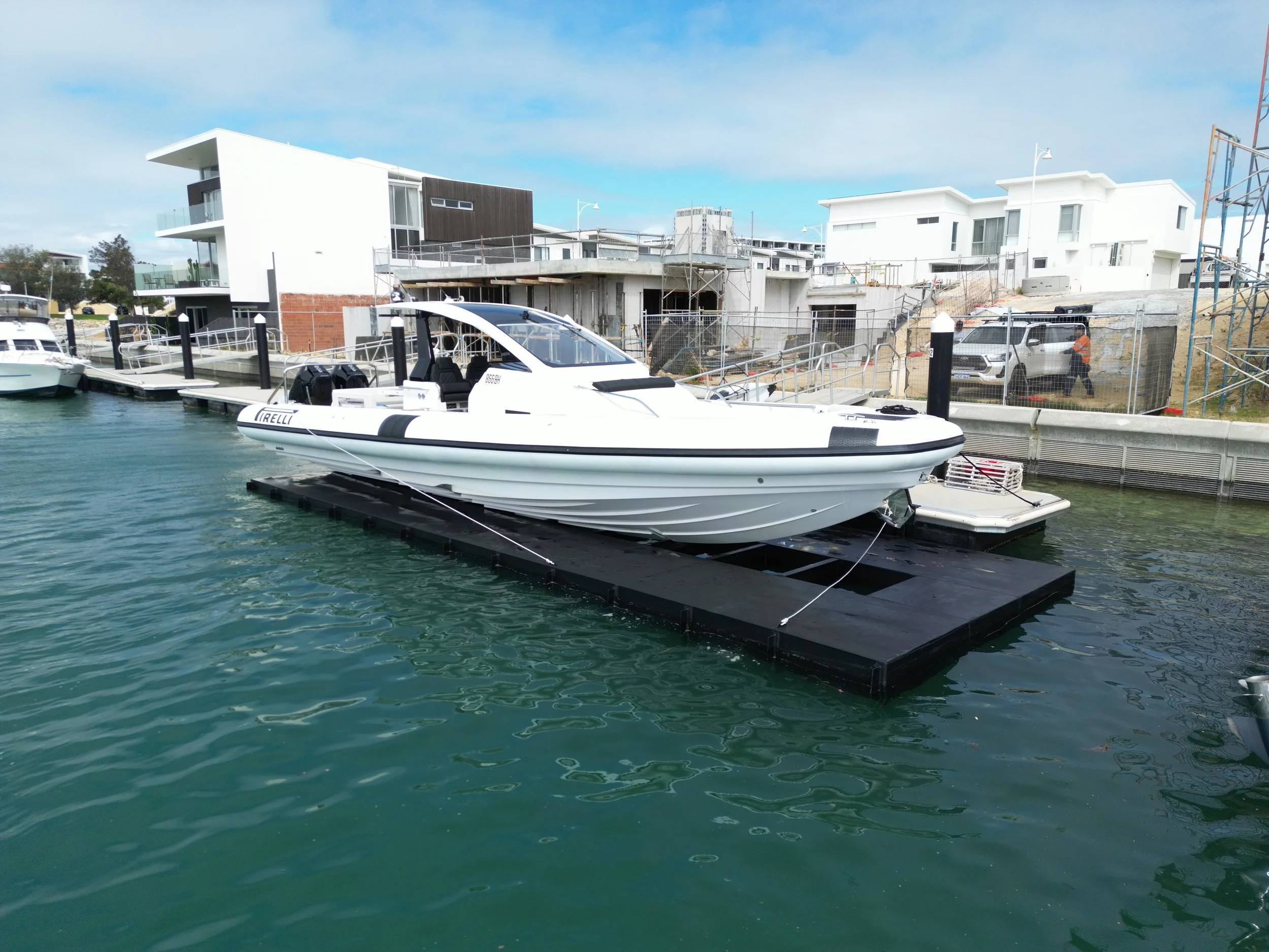 A white motorboat docked at a marina with modern white and black buildings under a partly cloudy sky in the background.