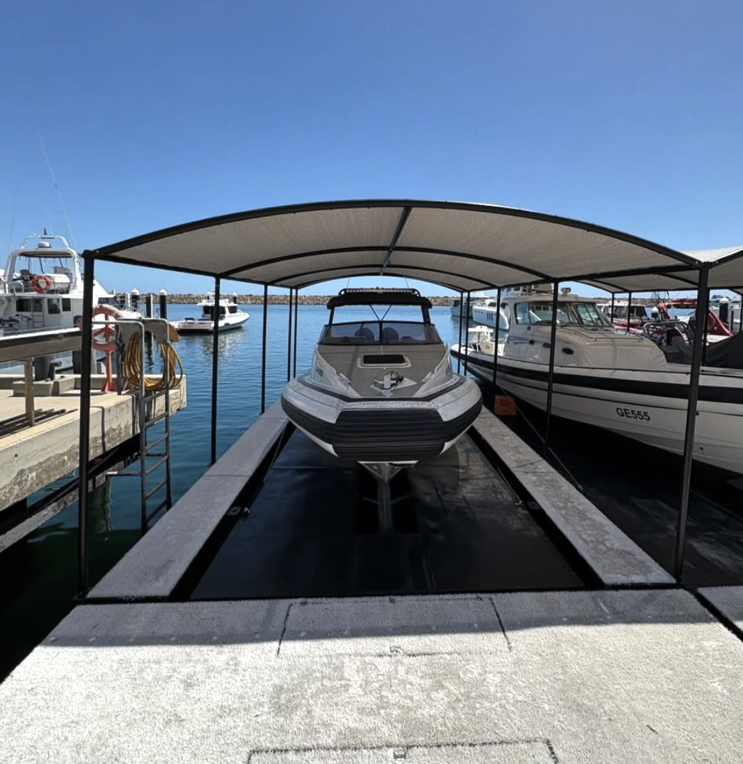 A motorboat under a covered boat slip at a marina, with a clear blue sky and calm water surrounding other boats docked nearby.