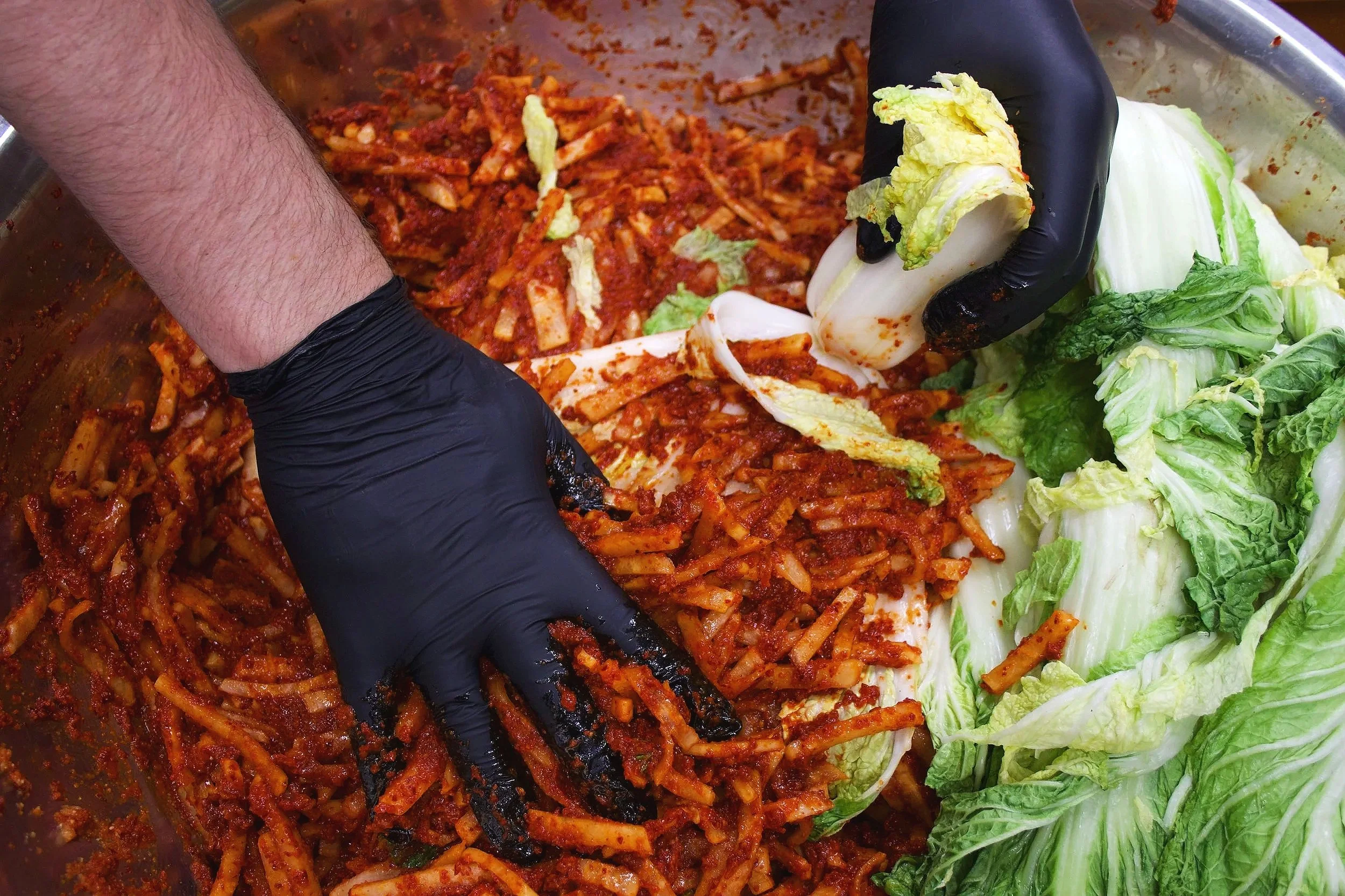 Matchstick cut daikons mixed with a red kimchi sauce being put on quarter cut napa cabbages by a person in black gloves