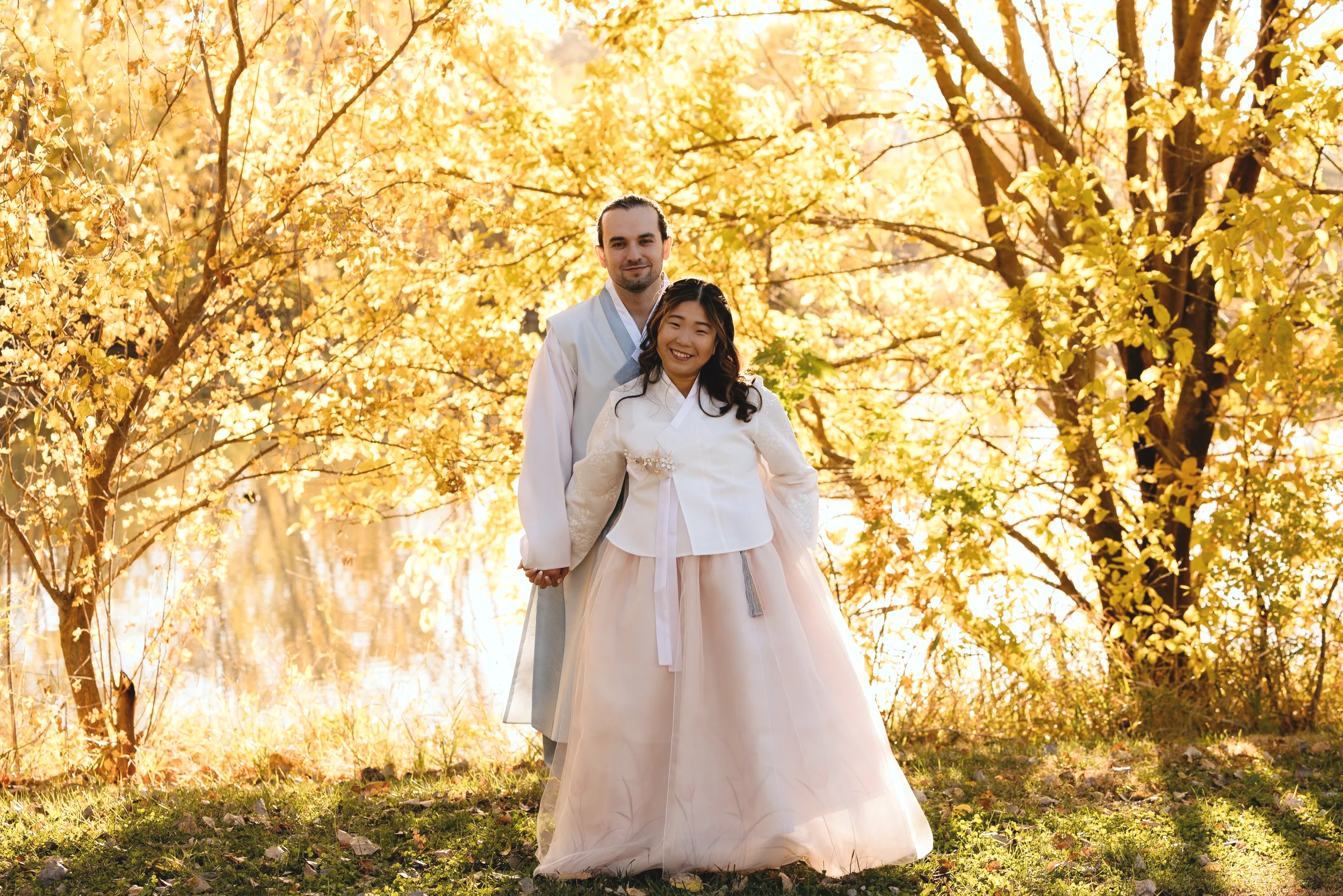 Two people (owners & founders of Sunmi Kimchi) in peach and blue color Hanbok (korean traditional wear). Tree and lake in the background with the sun shining behind the two people.