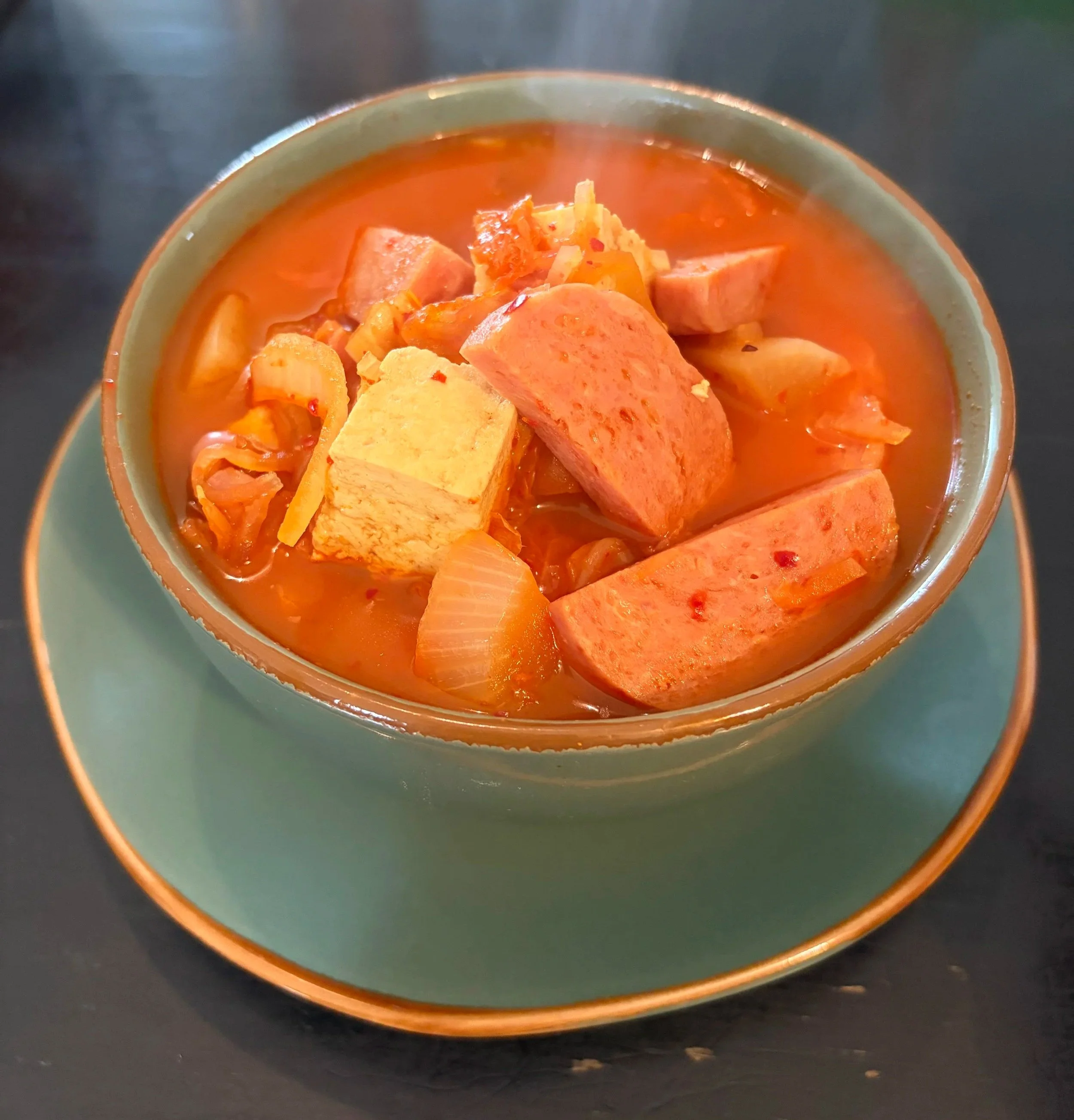 A green bowl on top of a green plate on a black table top. In the bowl is spam, tofu, onions, and kimchi.