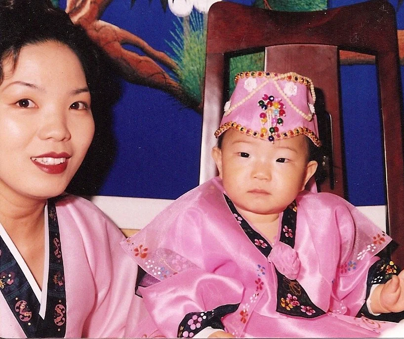 A woman with black hair wearing a pink hanbok (traditional korean wear) with a navy blue color and flower design. A toddler wear a pink hanbok with navy blue color with flower like embellishments and a headwear with colorful beads as decor