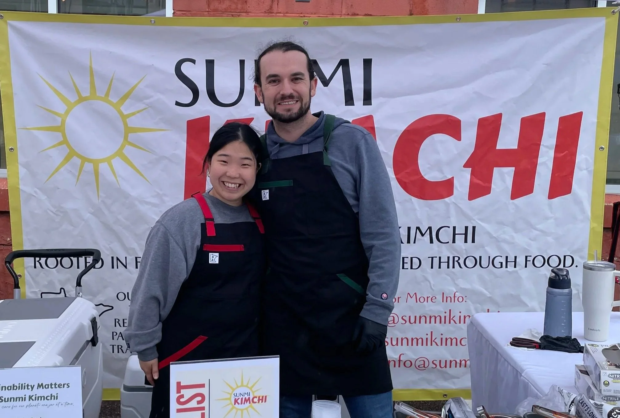 Two people in front of Sunmi Kimchi banner at farmers market