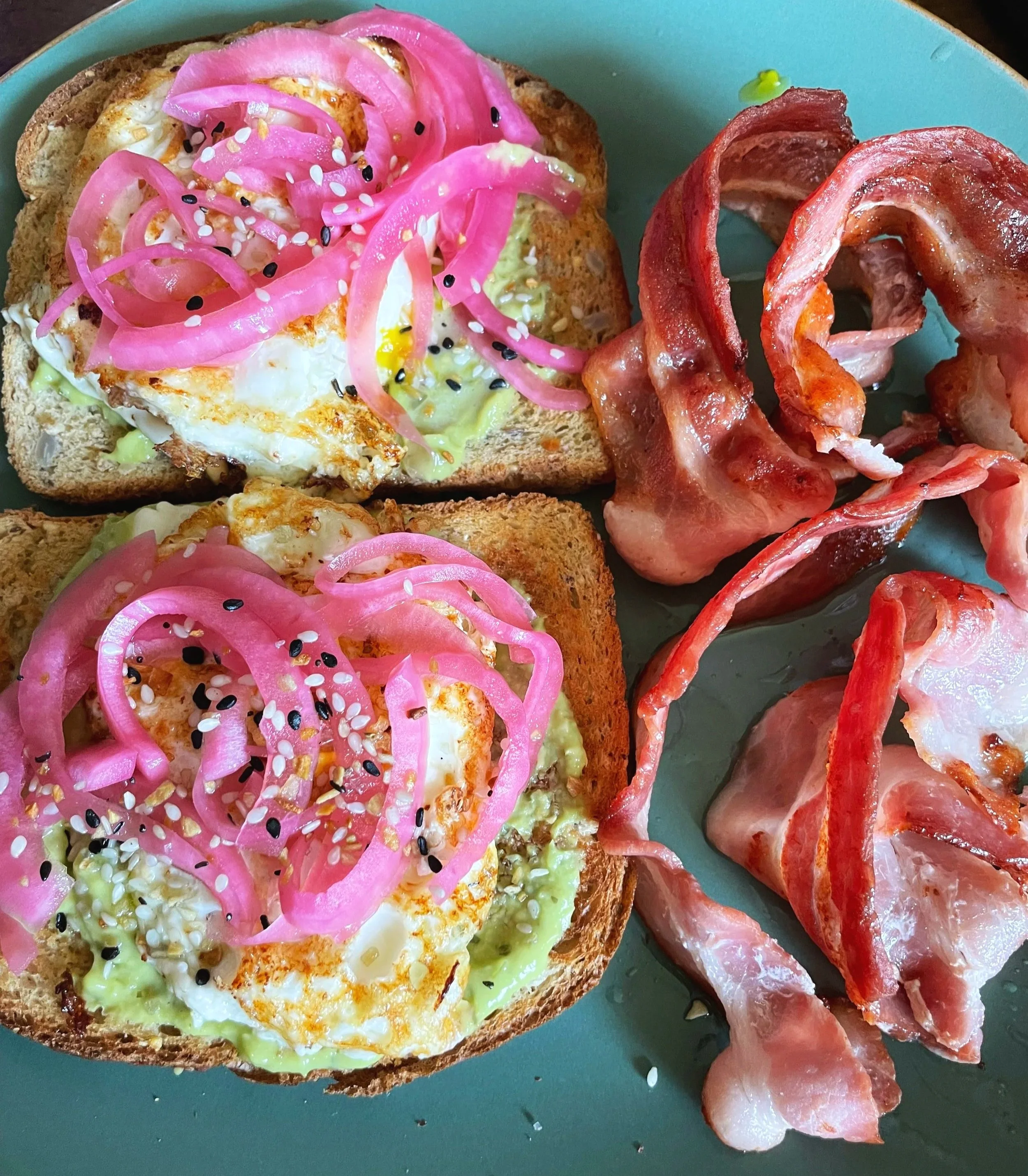 two slices of toast with avocado, eggs, pickled red onions, and everything bagel seasoning on top. It's plated on a green plate with 4 slices of bacon