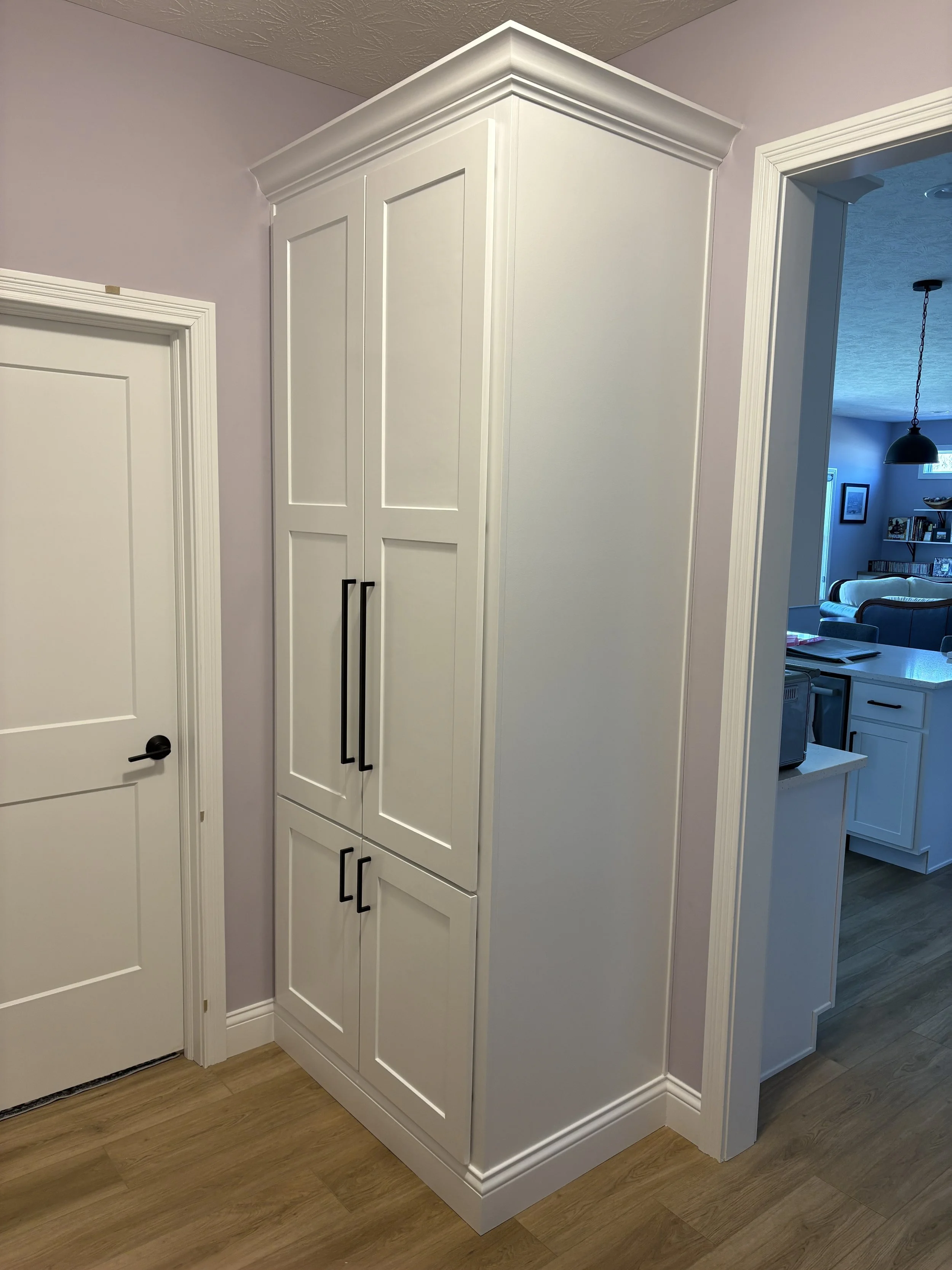 White painted built in armoire or wardrobe with black handles, crown, and baseboard in a mudroom with lvp floors, white doors, and lavender trim