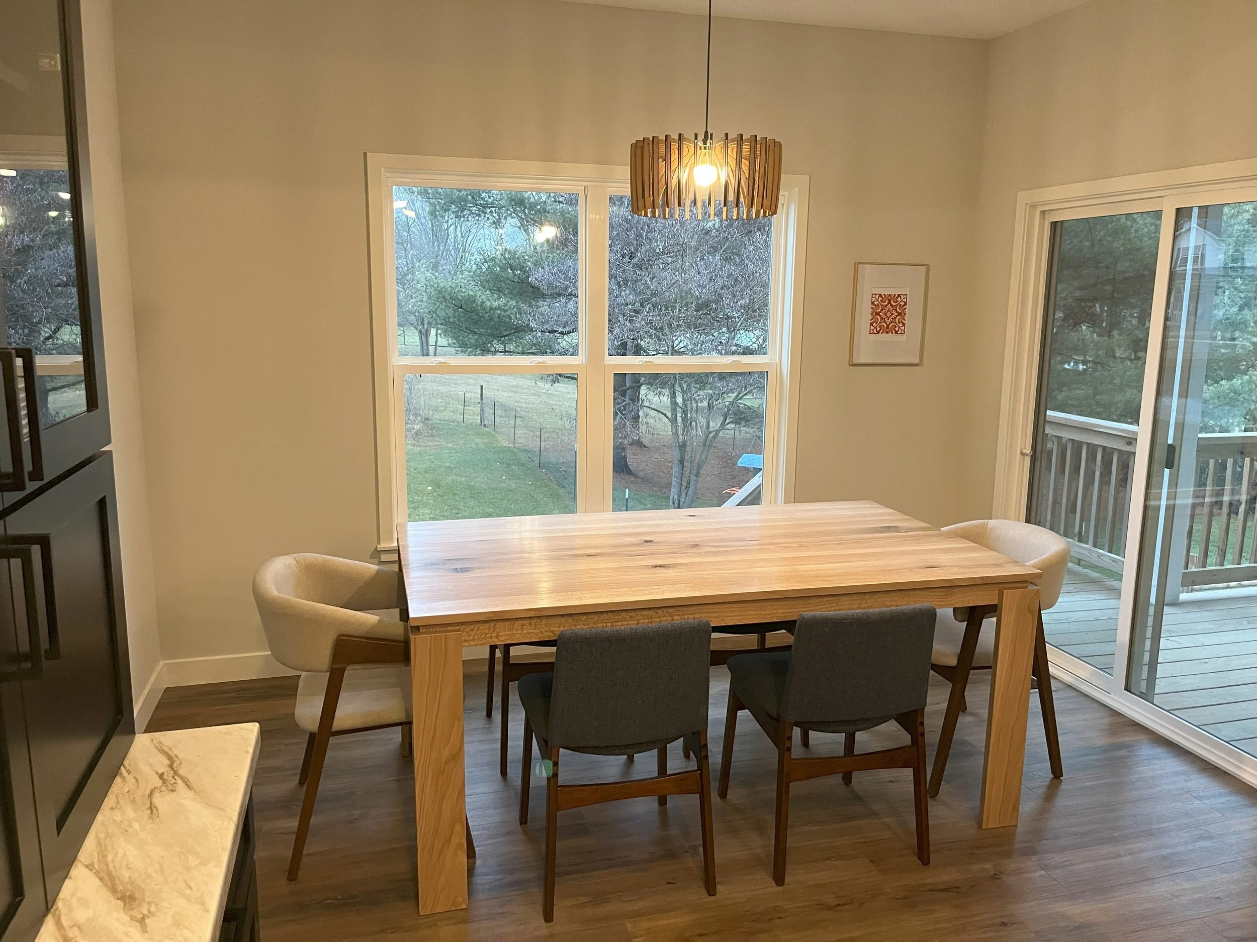 Dining room with a wooden table, six chairs, and a modern chandelier hanging above, large windows showing a backyard with trees and a sliding glass door leading to a deck.