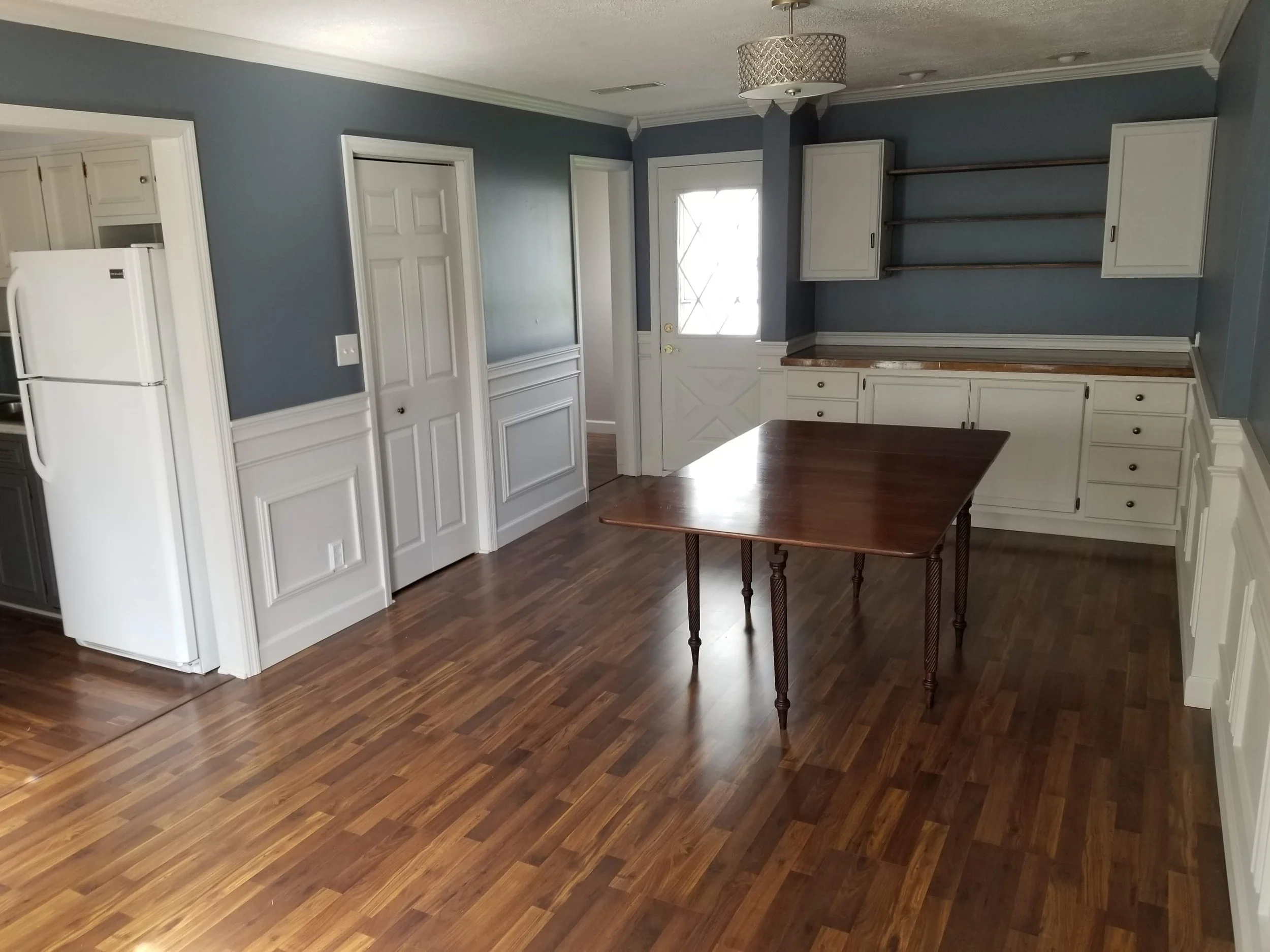 Empty kitchen with blue walls, wooden floor, white cabinets, a small wooden table in the center, and an exterior door with a small window. There are open shelves on the wall above the cabinets.