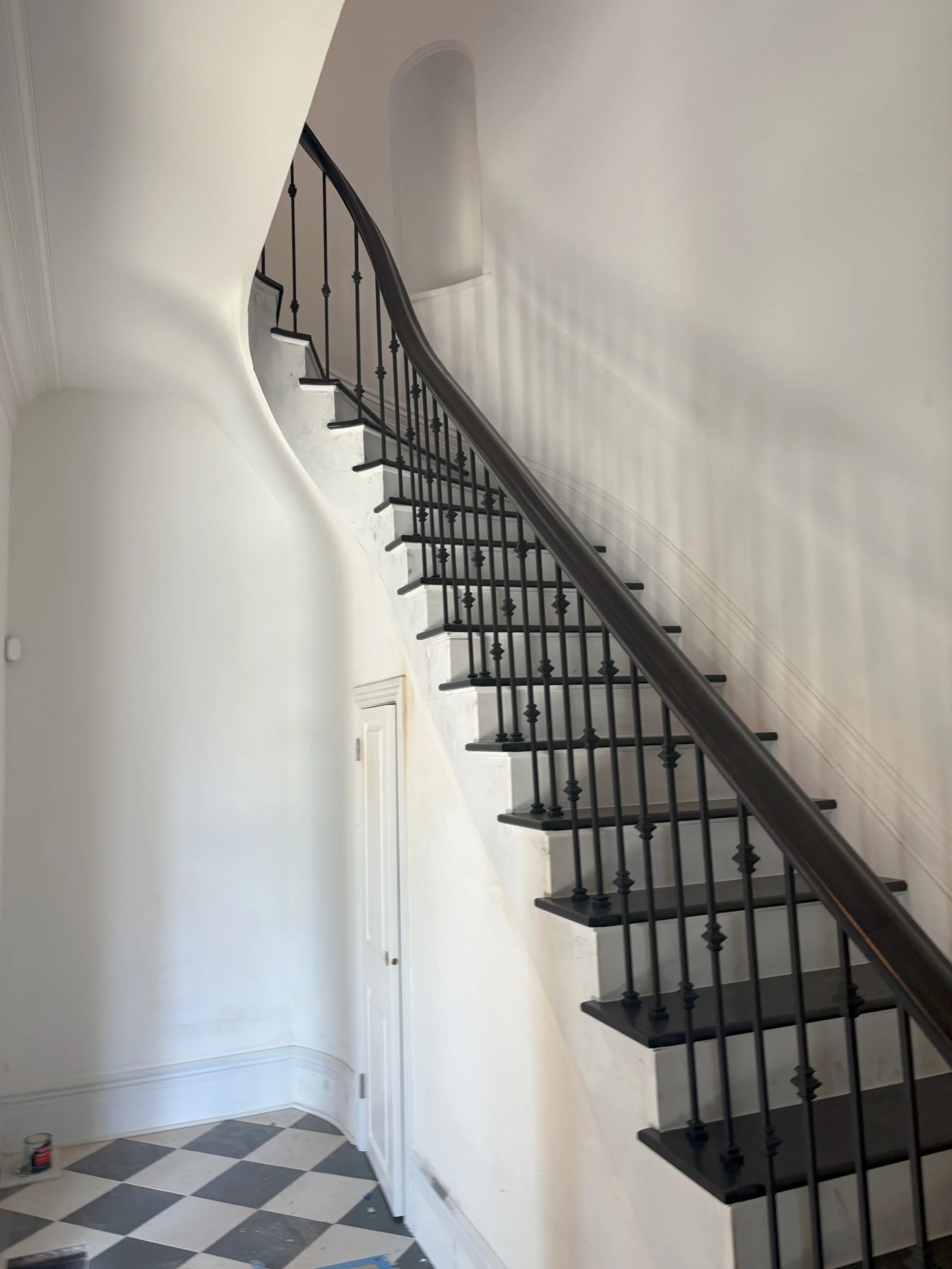 Interior view of a staircase with black handrails and white steps, leading upward with a twist. There is white wall on the side, with a small door underneath the staircase and a black and white checkered floor.