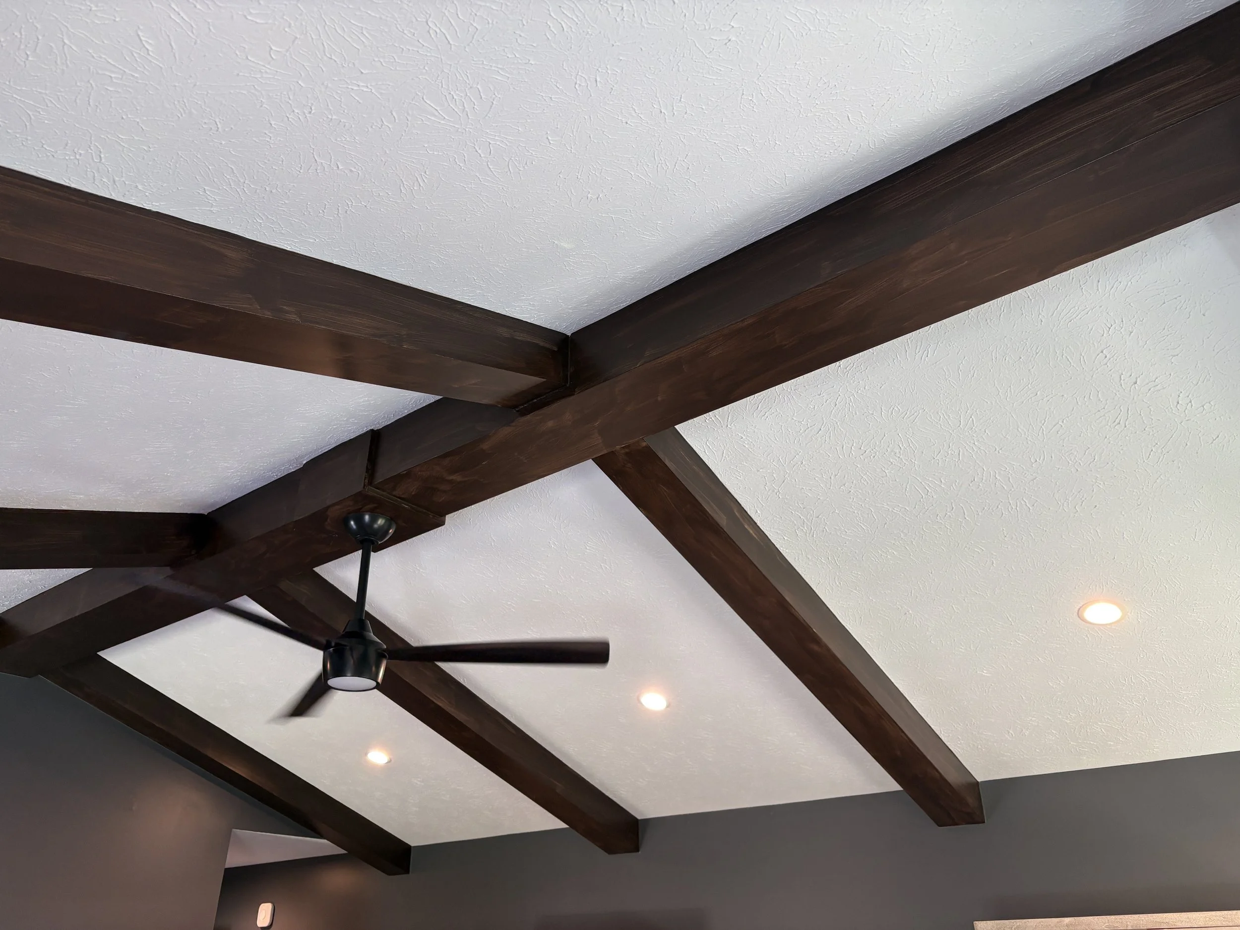 brown painted faux beams in a bedroom with a white ceiling, ceiling fan and gray/blue walls
