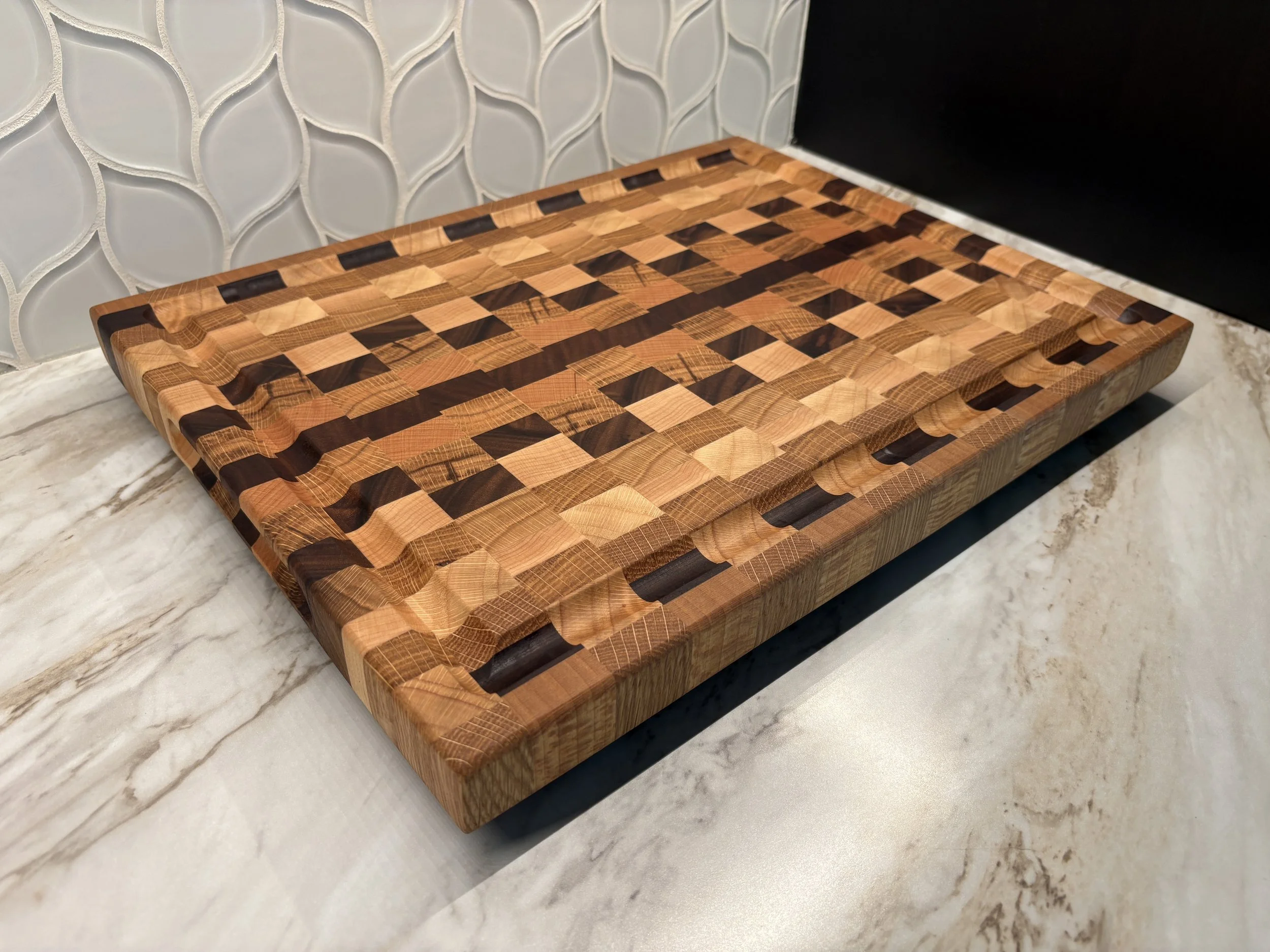 mixed hardwood cutting board with juice grooves on a white kitchen countertop with tiled backsplash and black cabinets, white oak, red oak, black oak, maple, walnut