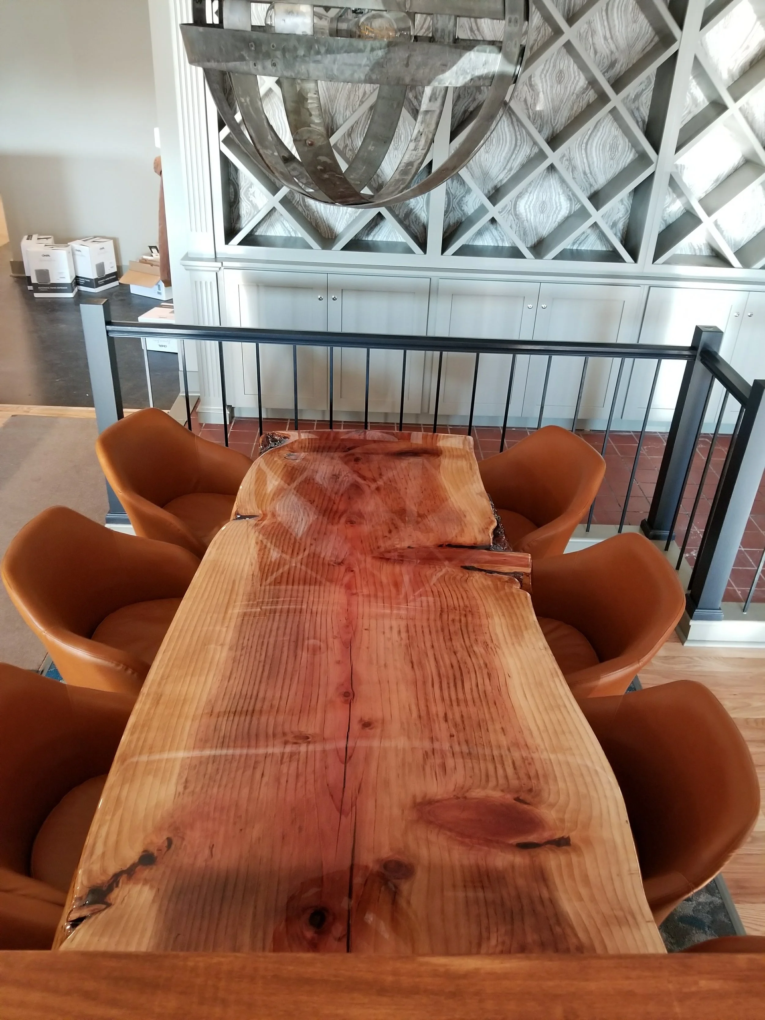 Top-down view of a rustic wooden dining table with a live edge, surrounded by six brown leather chairs, inside a modern room with decorative wall panels and a metal railing.