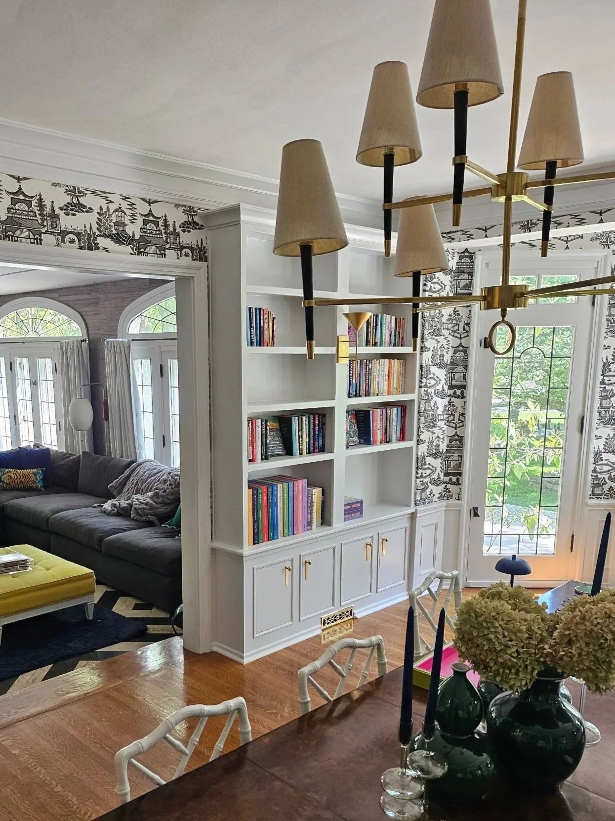 Interior view of a living room with white built-in bookshelf, hardwood floors, a dark sofa with pillows, a yellow ottoman, a dining table with black candlesticks, and a chandelier. Windows and glass doors allow natural light.