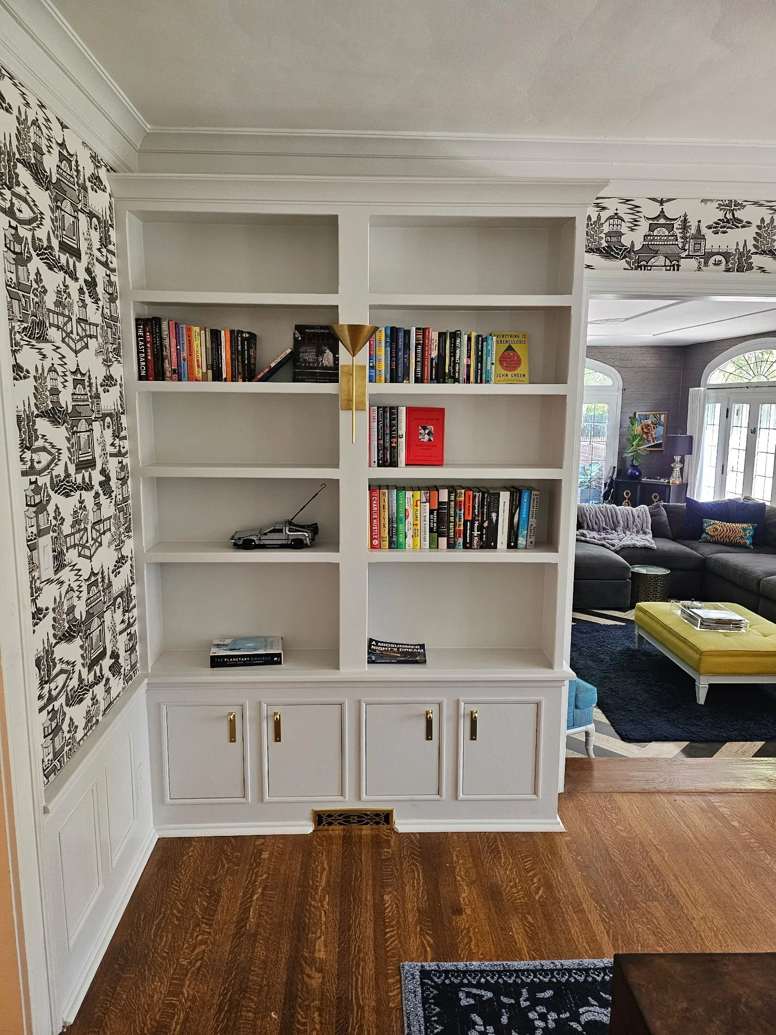 White built-in bookshelf with books and decorative items in a living room with patterned wallpaper and hardwood floor.