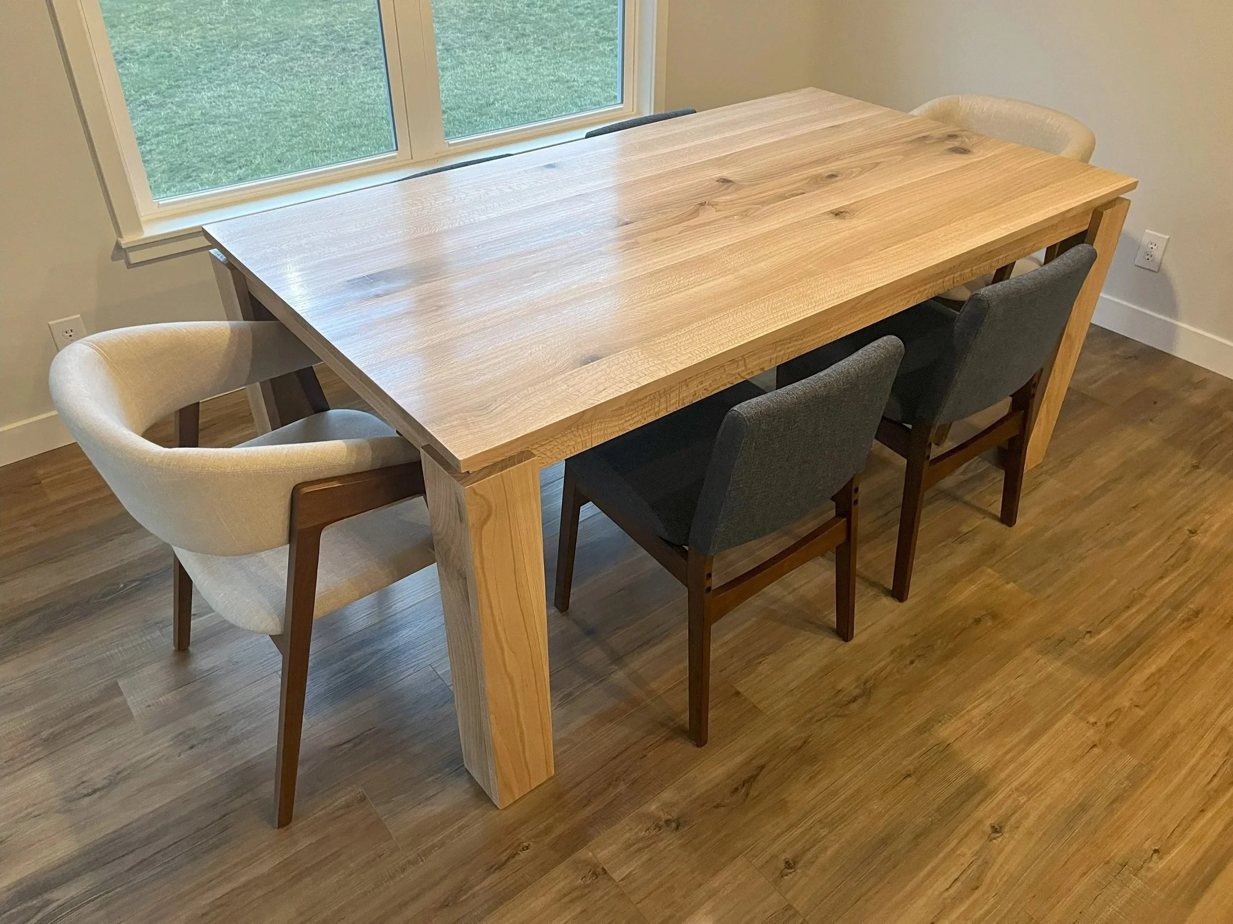 A wooden dining table with four chairs, two upholstered and two wooden, next to a large window showing green grass outside.