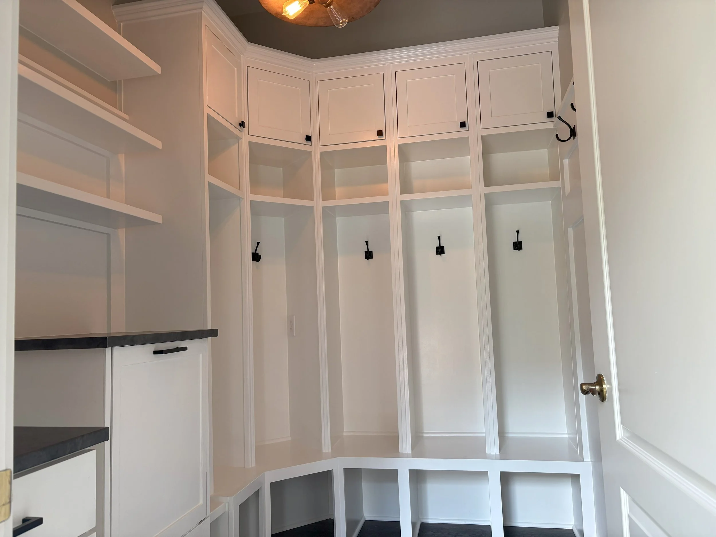 Empty built-in white closet with black hooks and shelves, black countertop, and a corner with cubbyholes, inside a room with a door and a ceiling light fixture.