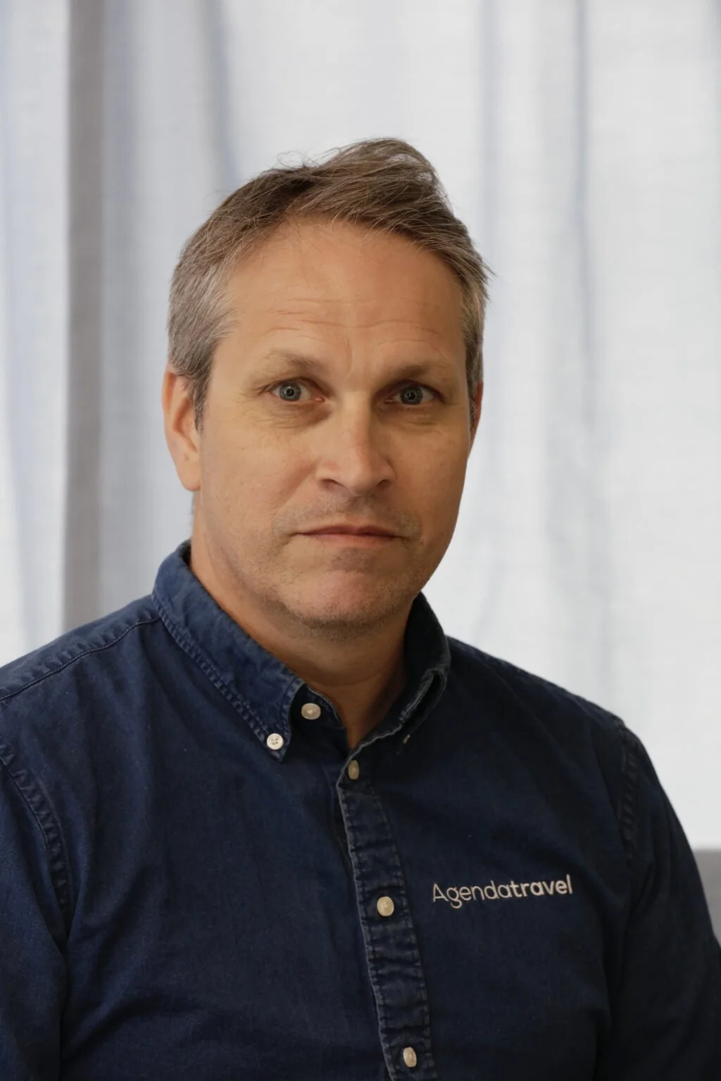 Man with gray hair wearing a dark blue shirt with 'Agendatravel' logo, standing against a light background.