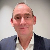 A man with a bald head and light skin, wearing a dark suit jacket and a white shirt, smiling in front of a plain, light-colored background.