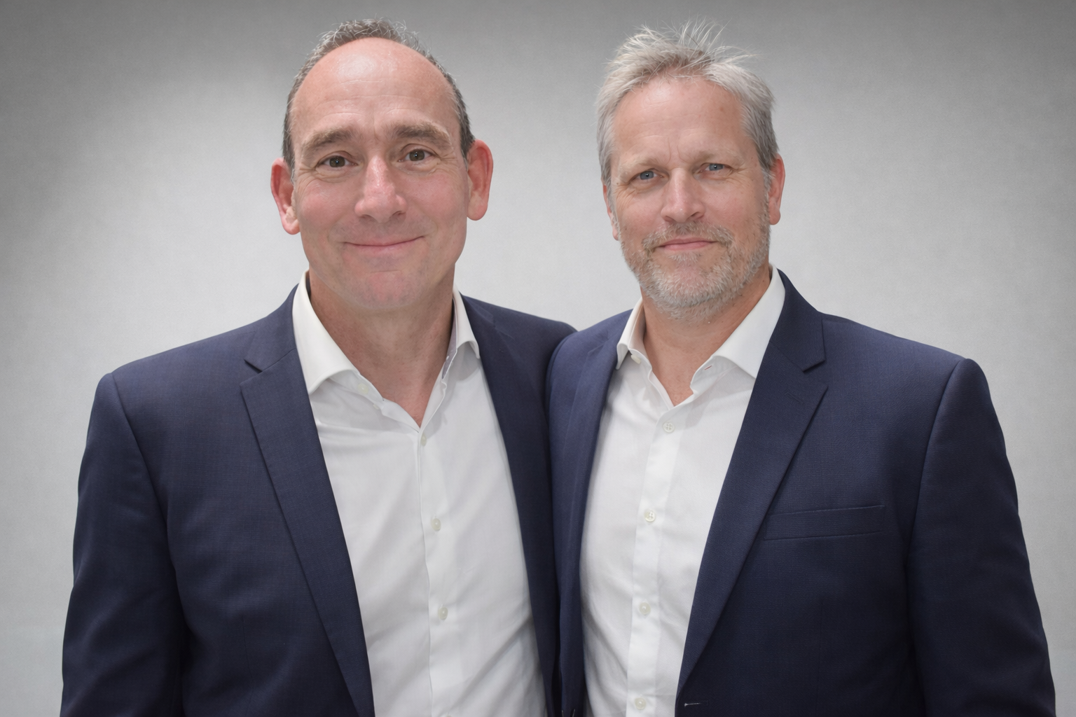 Two middle-aged men in suits standing together and smiling at the camera against a plain background.