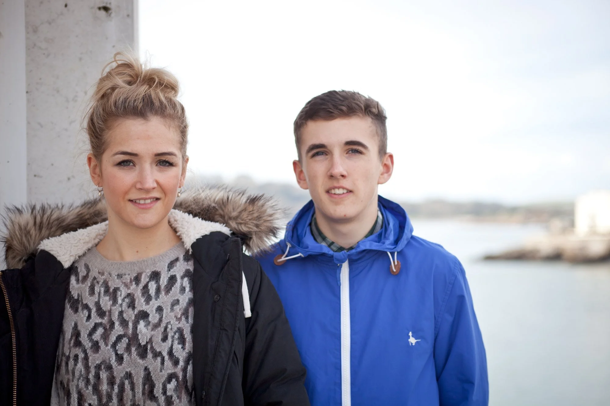 A young woman and a young man standing outdoors near water, both looking at the camera. The woman is wearing a black jacket with a fur-lined hood and a leopard print sweater. The man is wearing a bright blue jacket with a plaid shirt underneath.