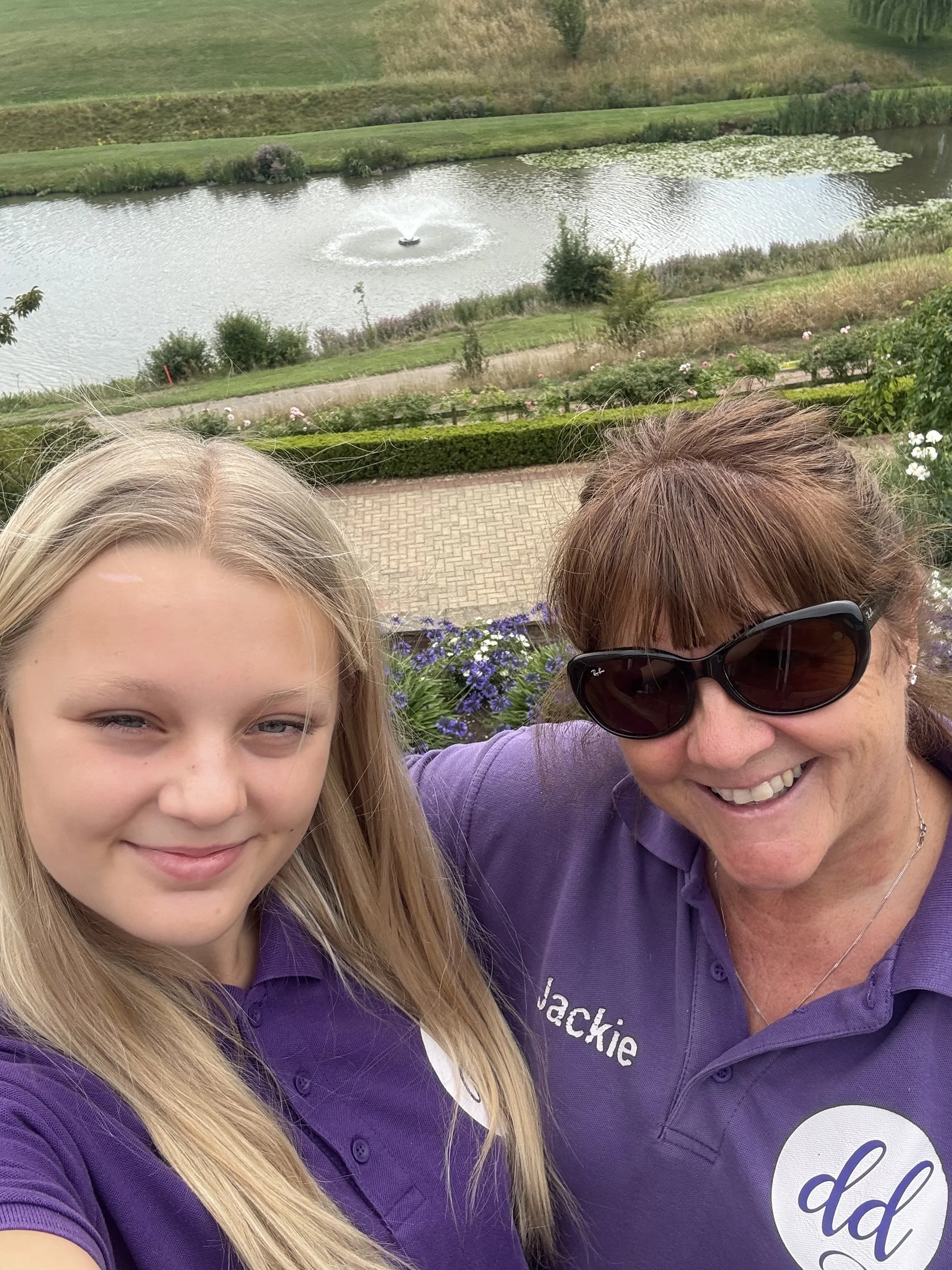 Two women taking a selfie with a pond featuring a fountain in the background. One woman is young with long blonde hair, and the other is older with brown hair, wearing sunglasses. Both are smiling and wearing purple shirts.