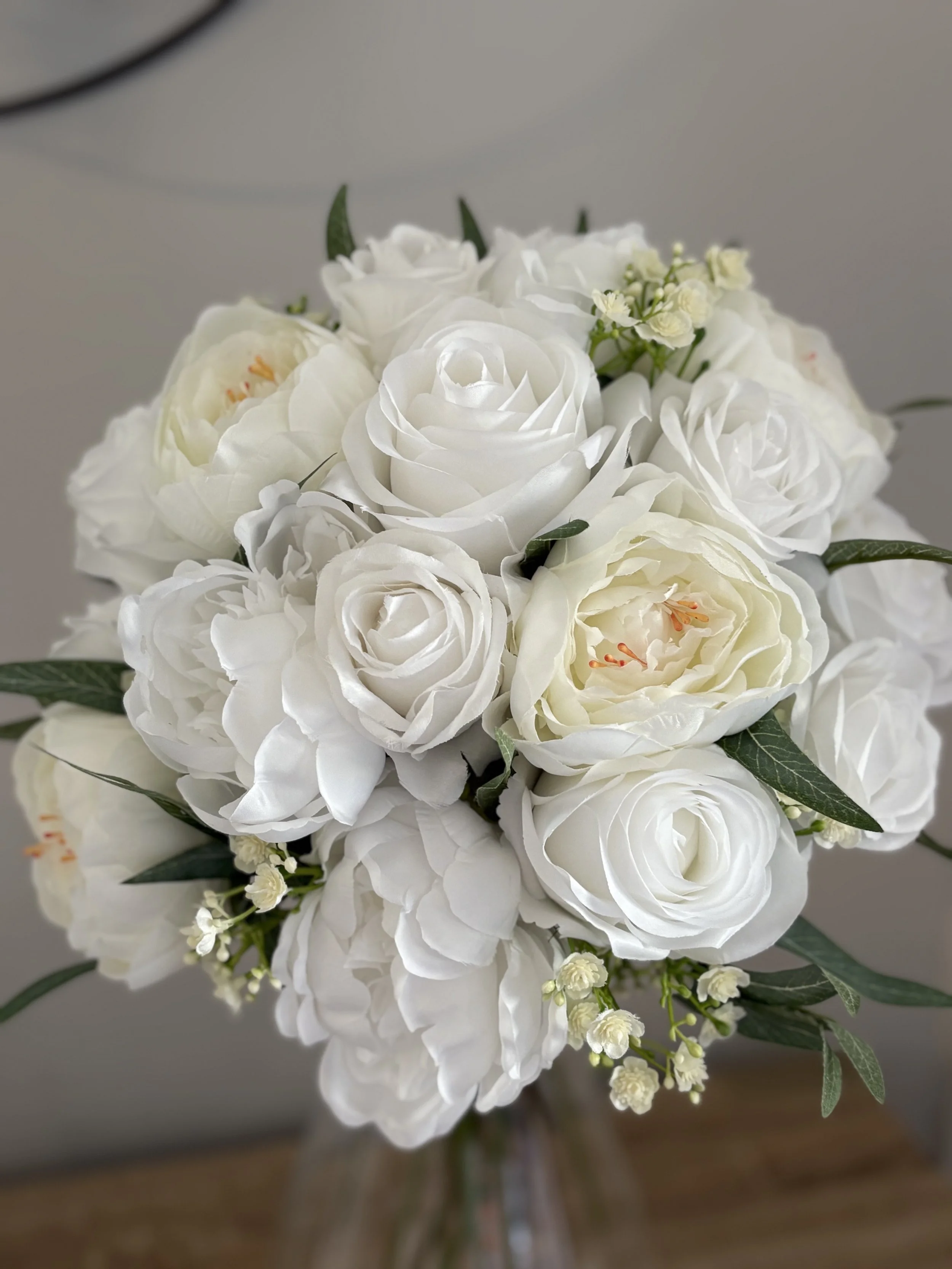A bouquet of white roses, peonies, and small white flowers with green leaves in a glass vase.