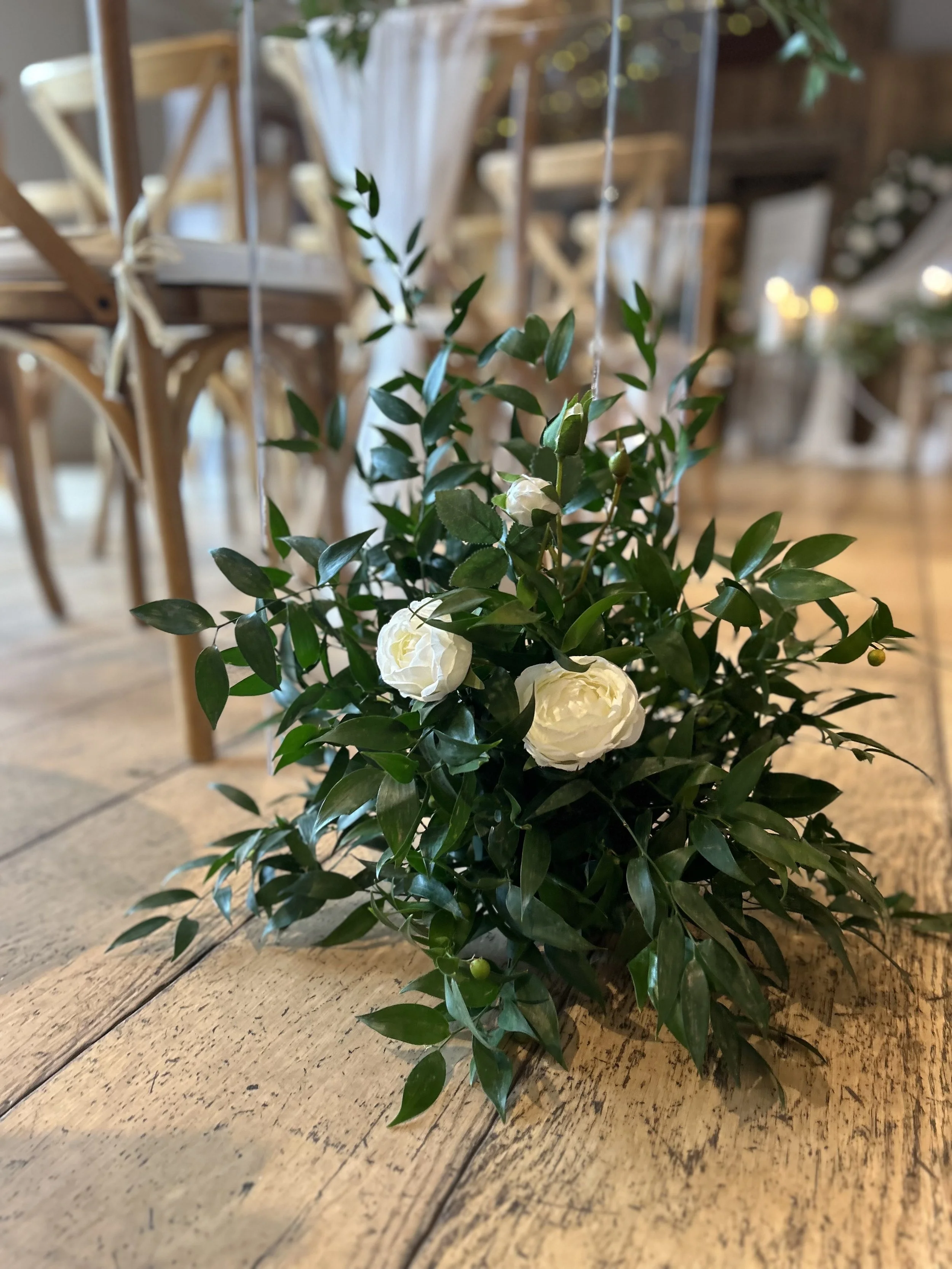 Small floral arrangement with white roses and green leaves on a wooden table in a rustic setting.