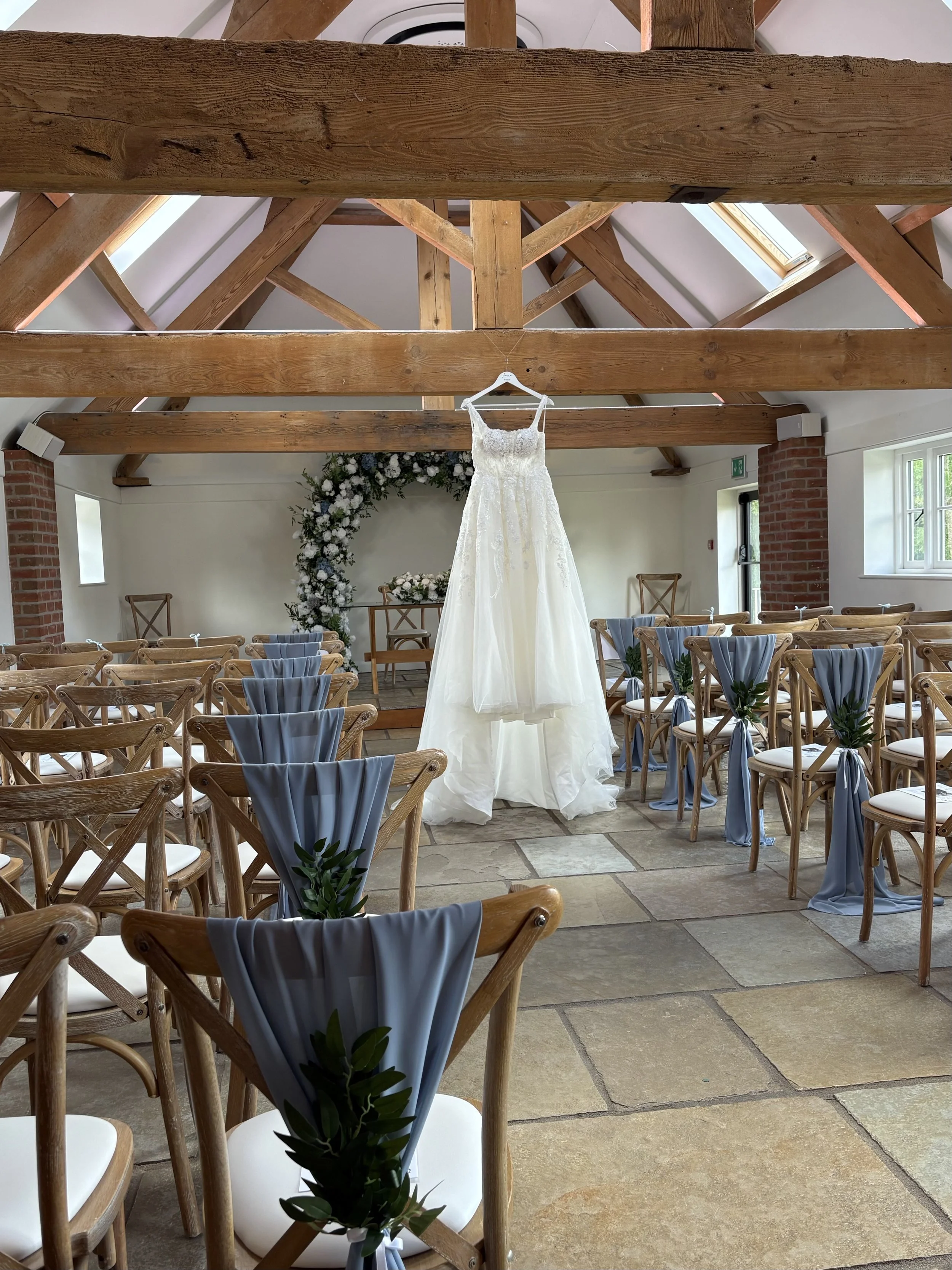 A wedding dress hanging from a wooden beam inside a decorated wedding ceremony venue, with wooden chairs adorned with gray fabric and green leaves, and a floral archway in the background.