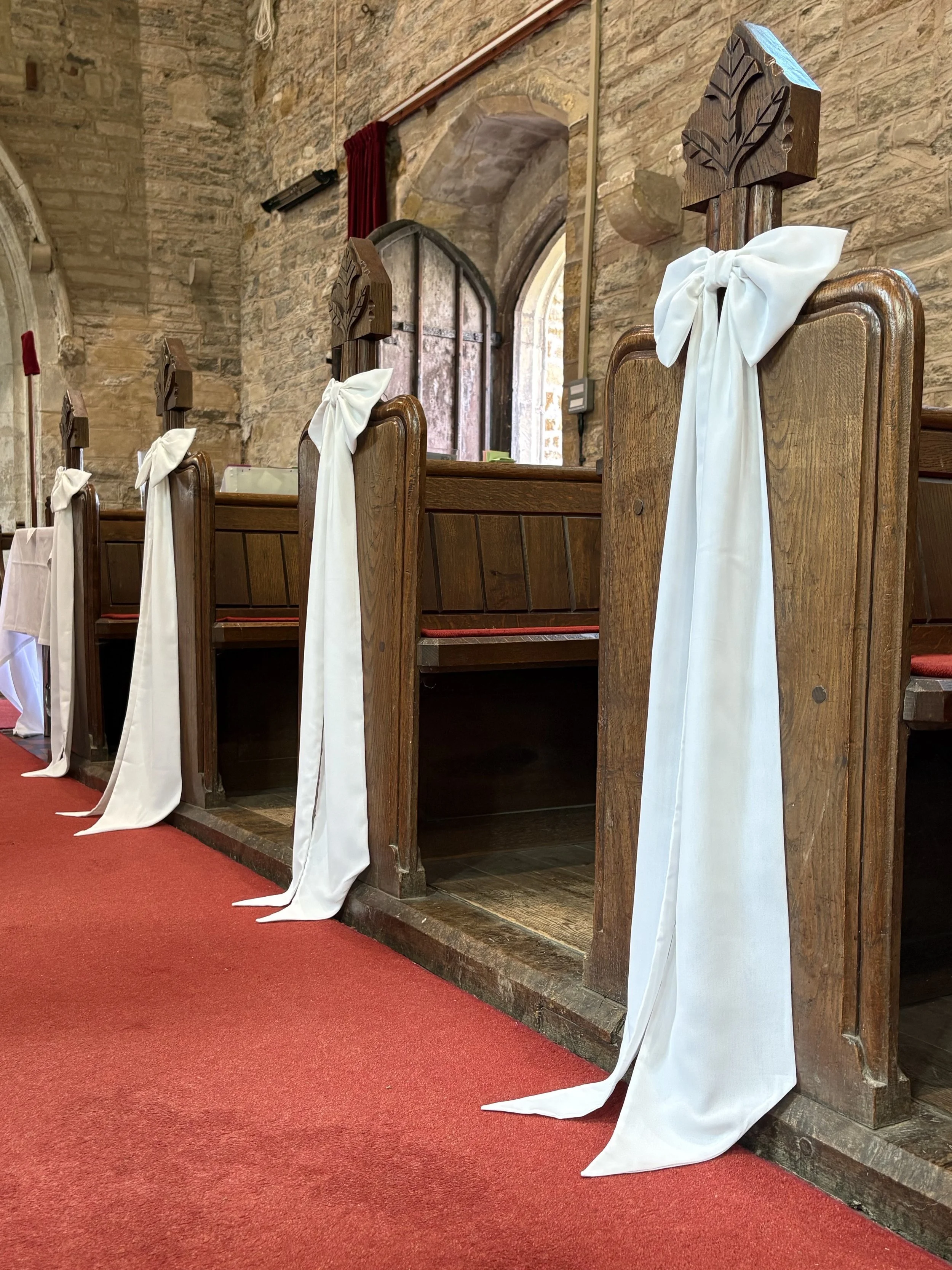 Wooden pews decorated with white fabric bows inside a stone chapel with arched windows and a red carpet aisle.