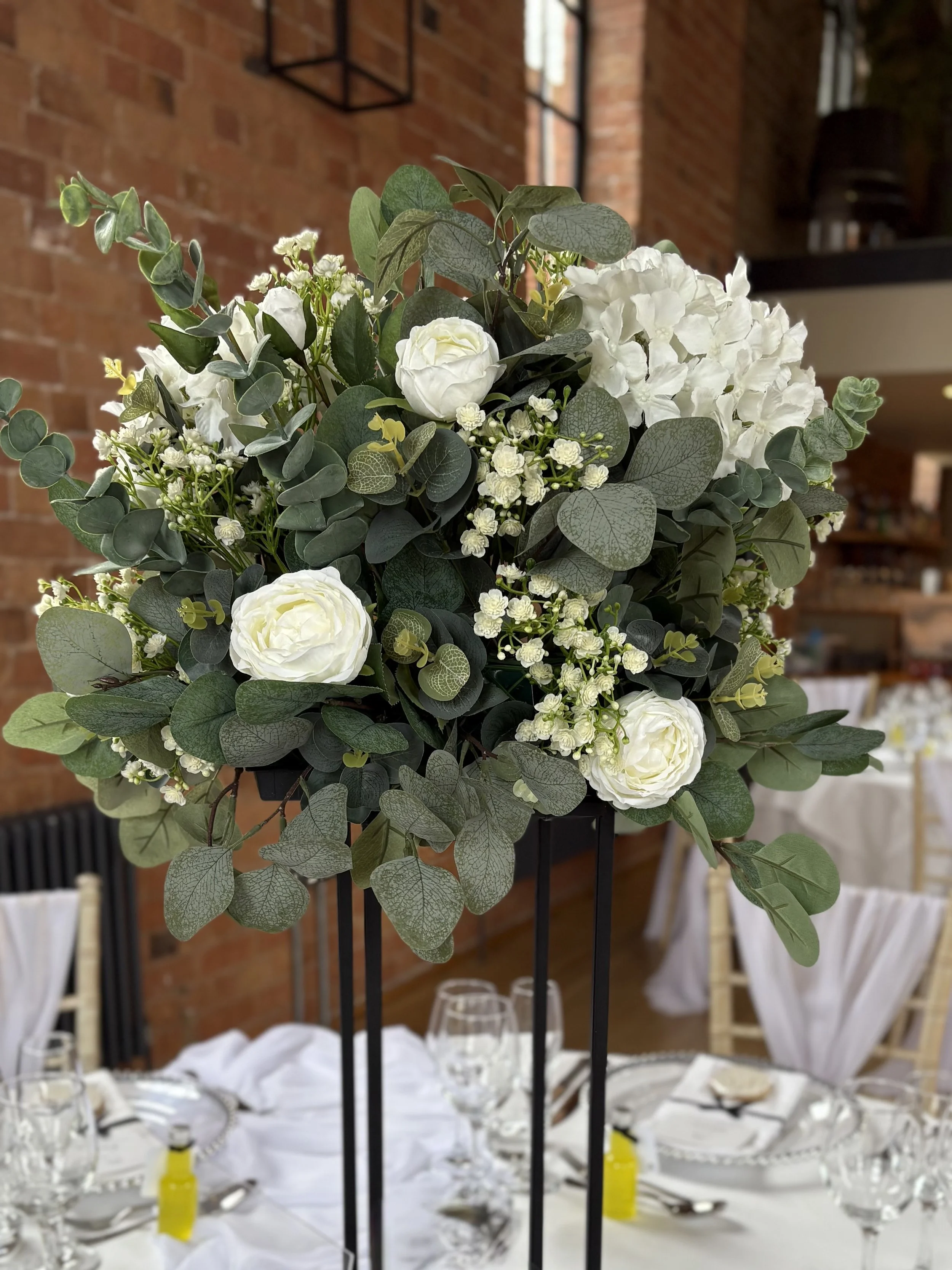 A floral centerpiece with white roses, hydrangeas, and greenery on a tall black stand, set on a white tablecloth at a decorated event with glassware and chairs.