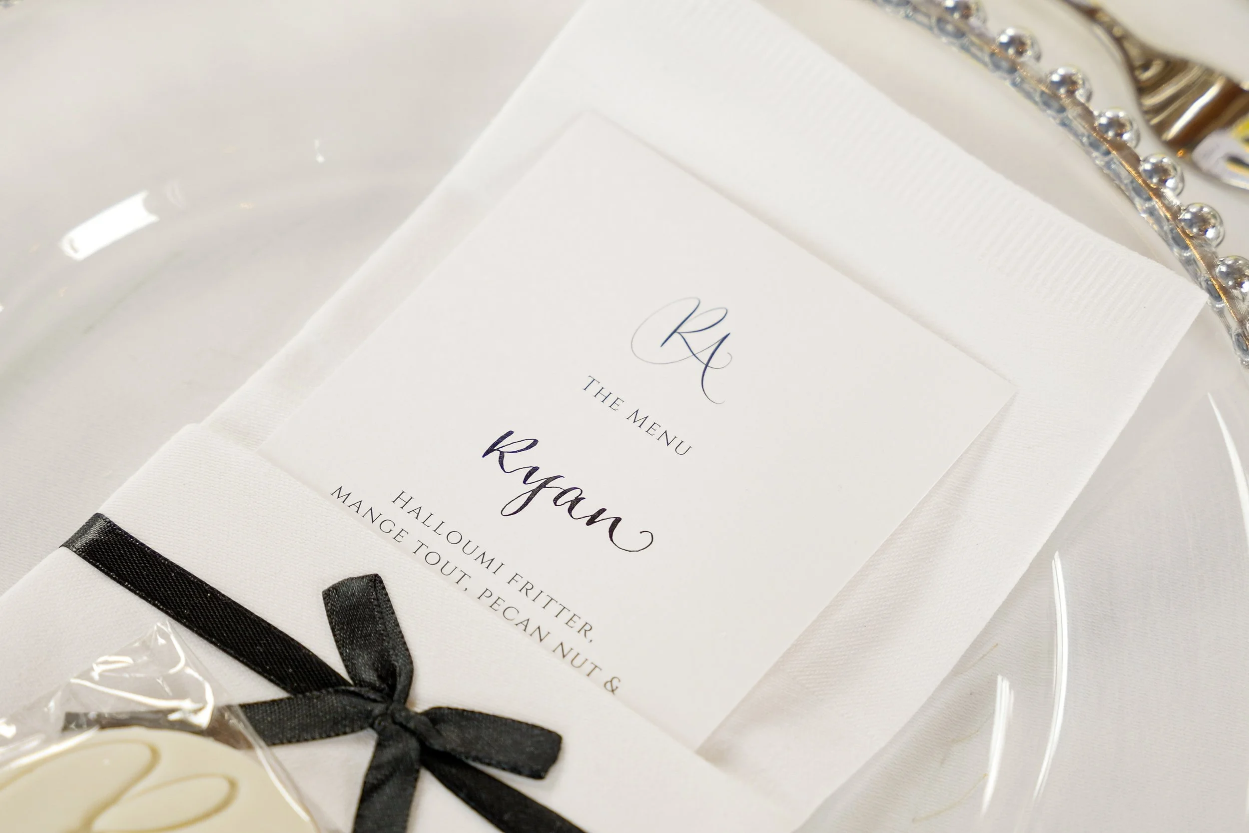 Elegant white place card with black ribbon, displaying a menu for a wedding or formal event, featuring the name Ryan and a stylized initials R and C at the top.