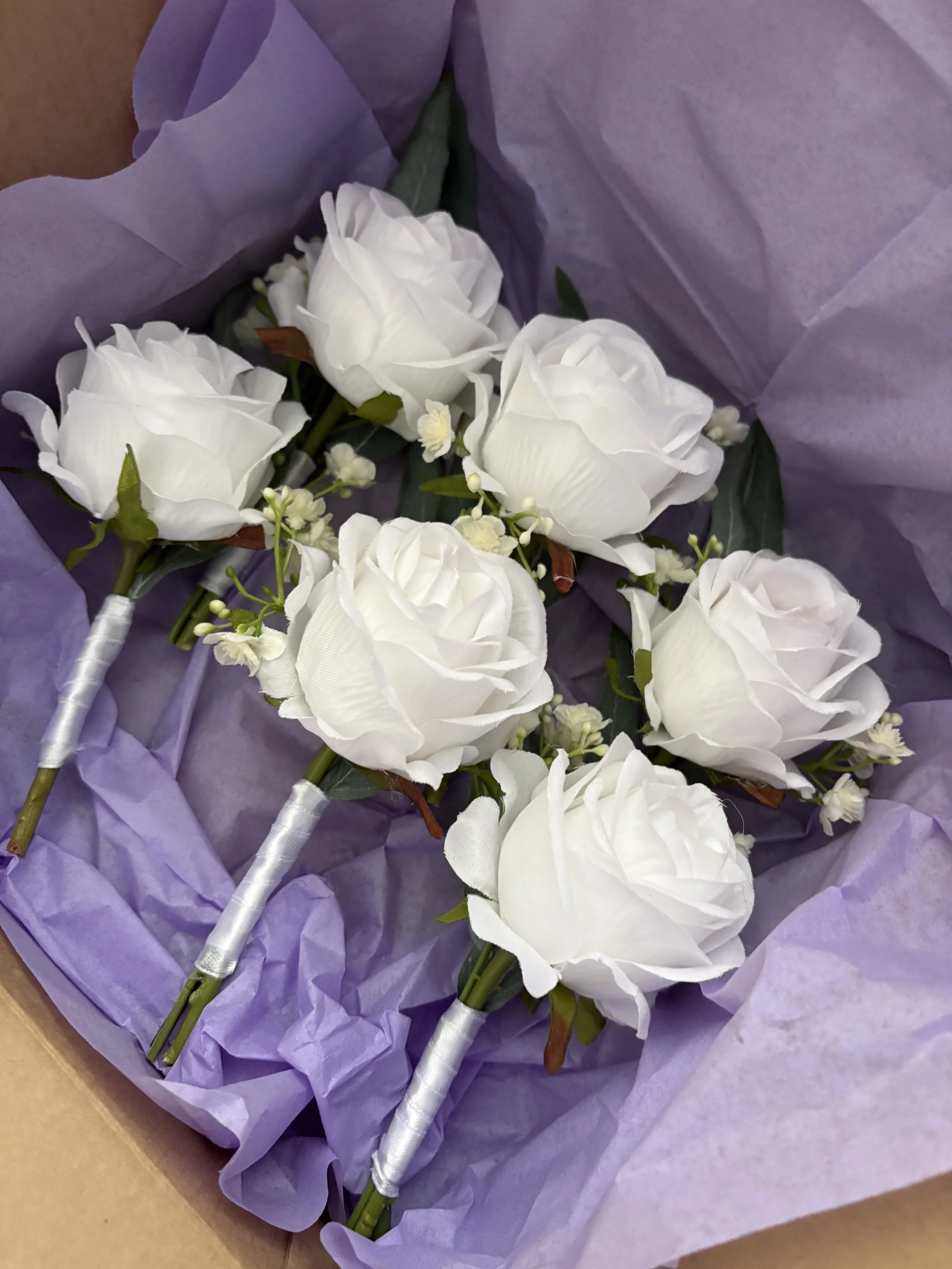 Six white roses arranged with small filler flowers and greenery, wrapped in purple tissue paper, inside a brown box.