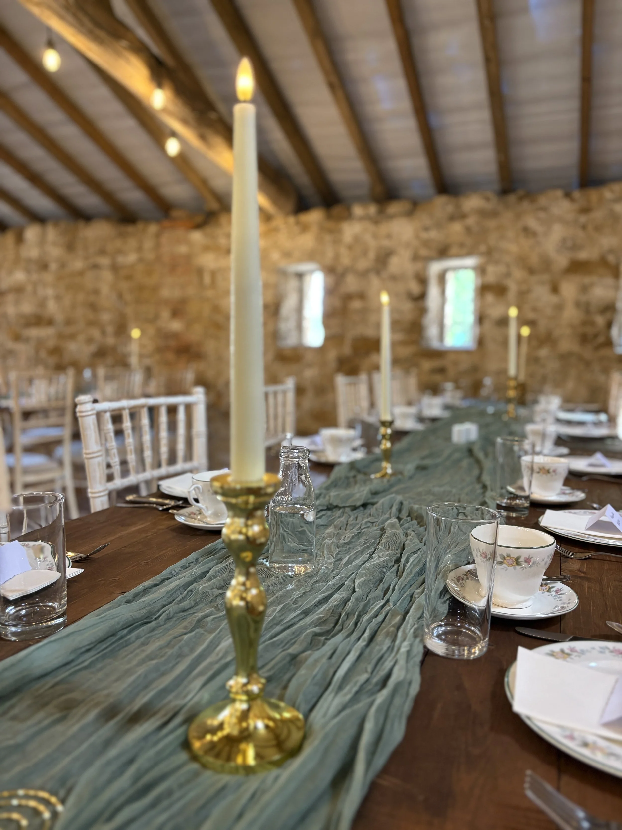 Wedding reception table with a green fabric table runner, gold candle holders with tall white candles, teacups, glass water bottles, and place settings in a room with stone walls and wooden beams.