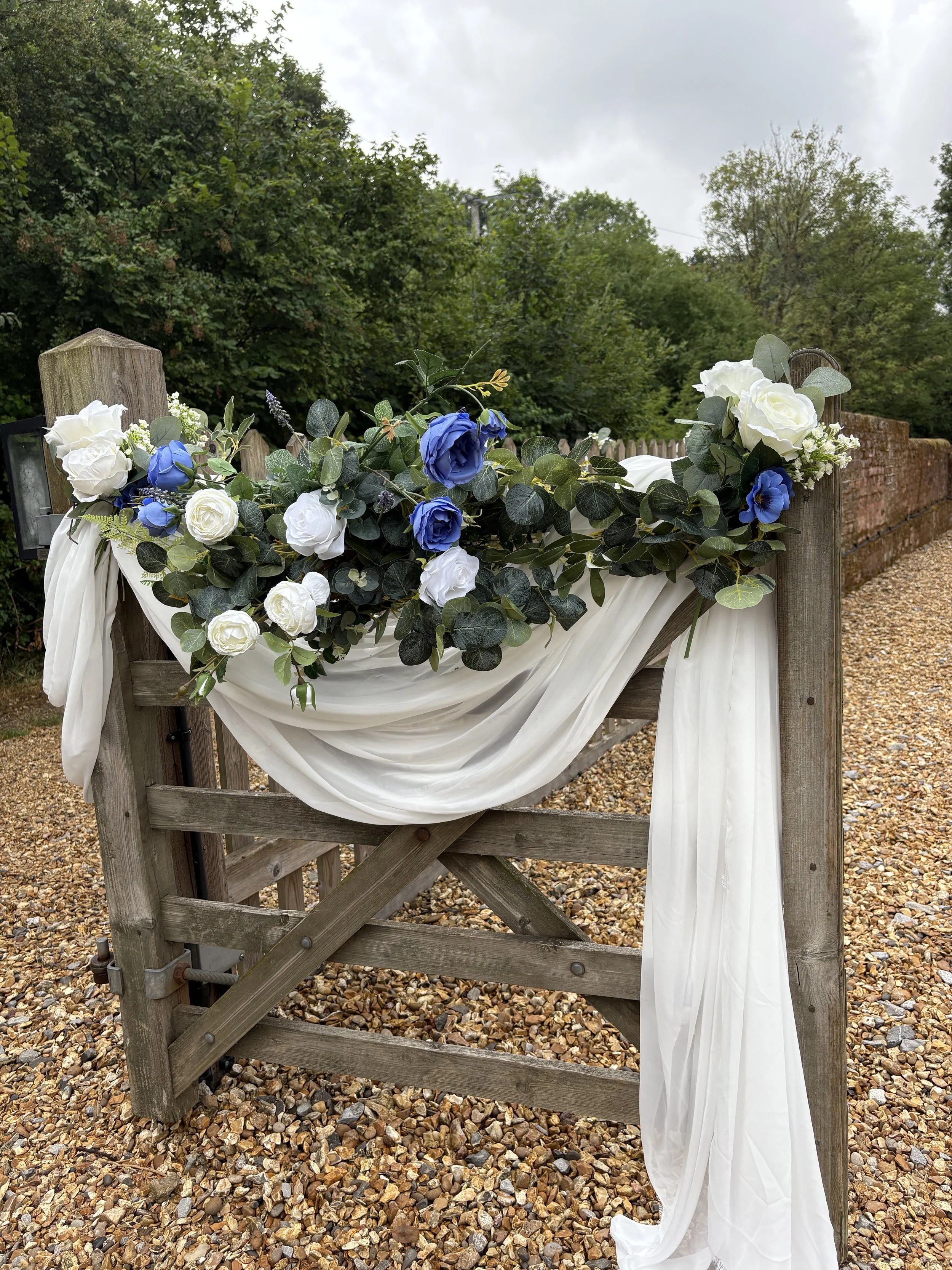 Wooden gate decorated with white and blue flowers, green leaves, and white fabric, outdoors with trees and a cloudy sky in the background.
