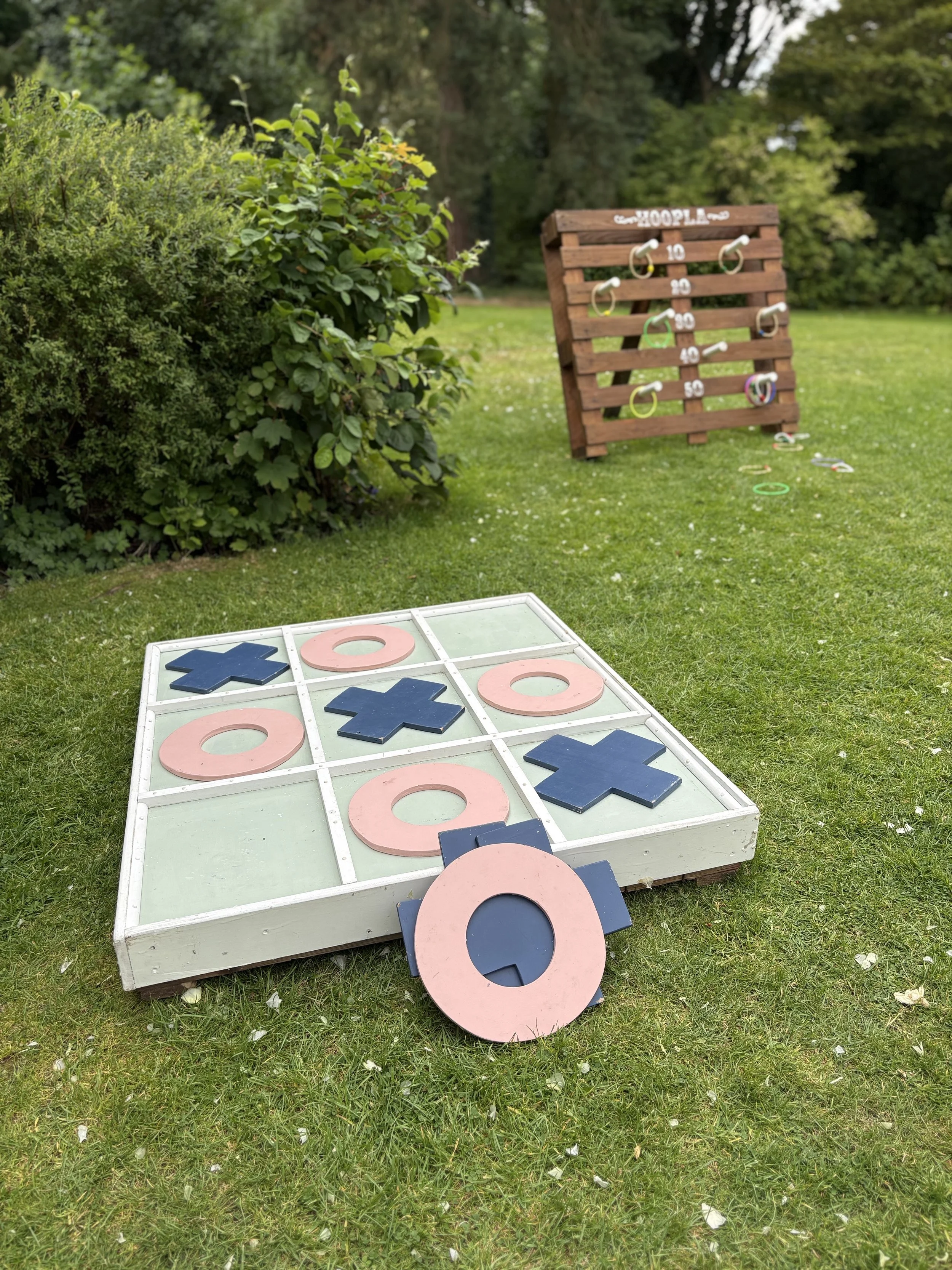 Outdoor yard game of tic-tac-toe with large wooden X and O pieces on a glass board, and a giant ring toss game on a wooden stand in the background.