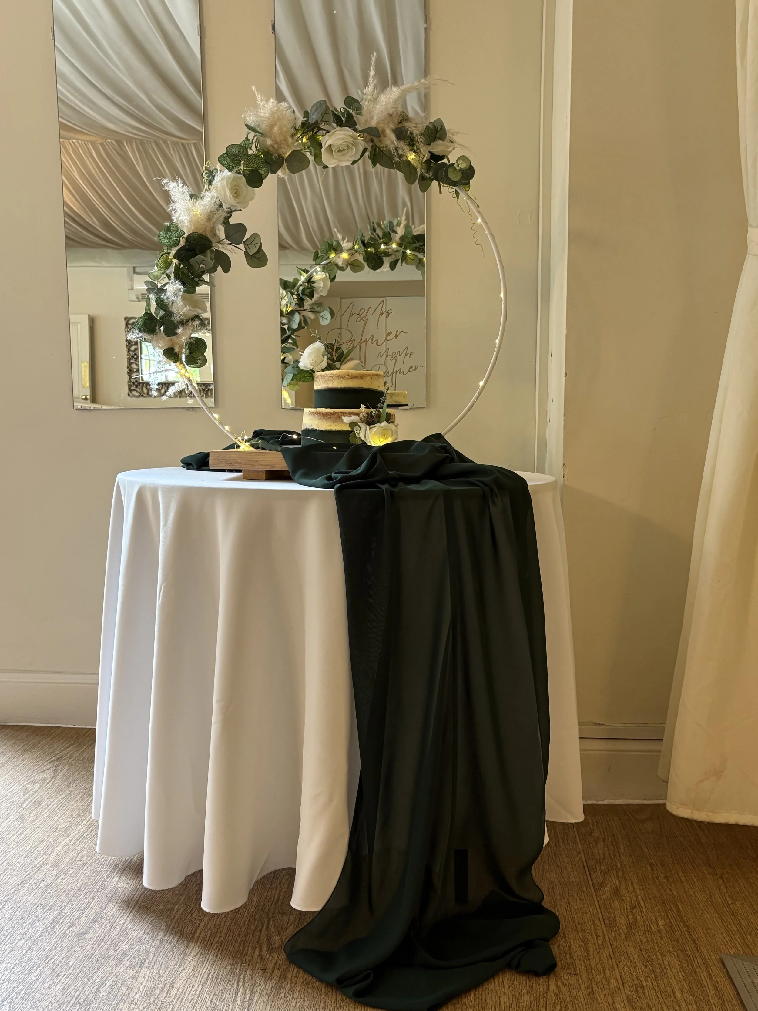 Decorative wedding cake on a round table with a black draped cloth, set in front of a crown-shaped floral arch, with a mirror and framed artwork in the background.